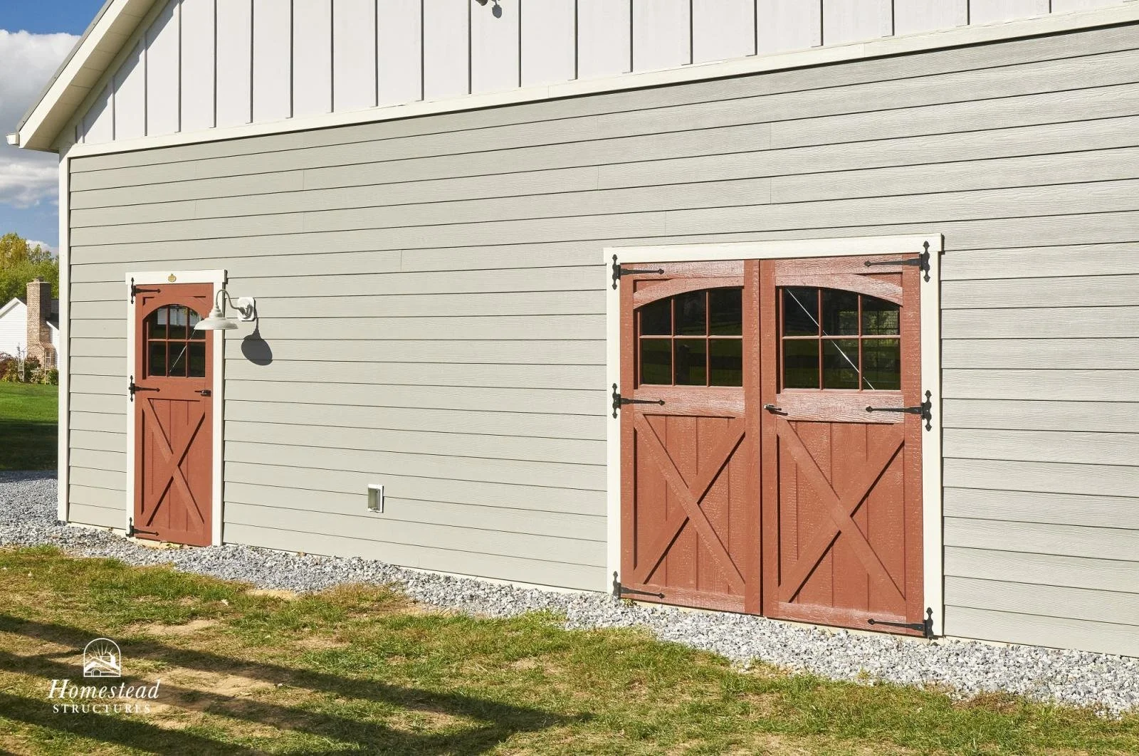 Exterior of a gray barn with a small door and large double doors, both with red wood finish and windowpanes, on a grassy yard with gravel around the base.