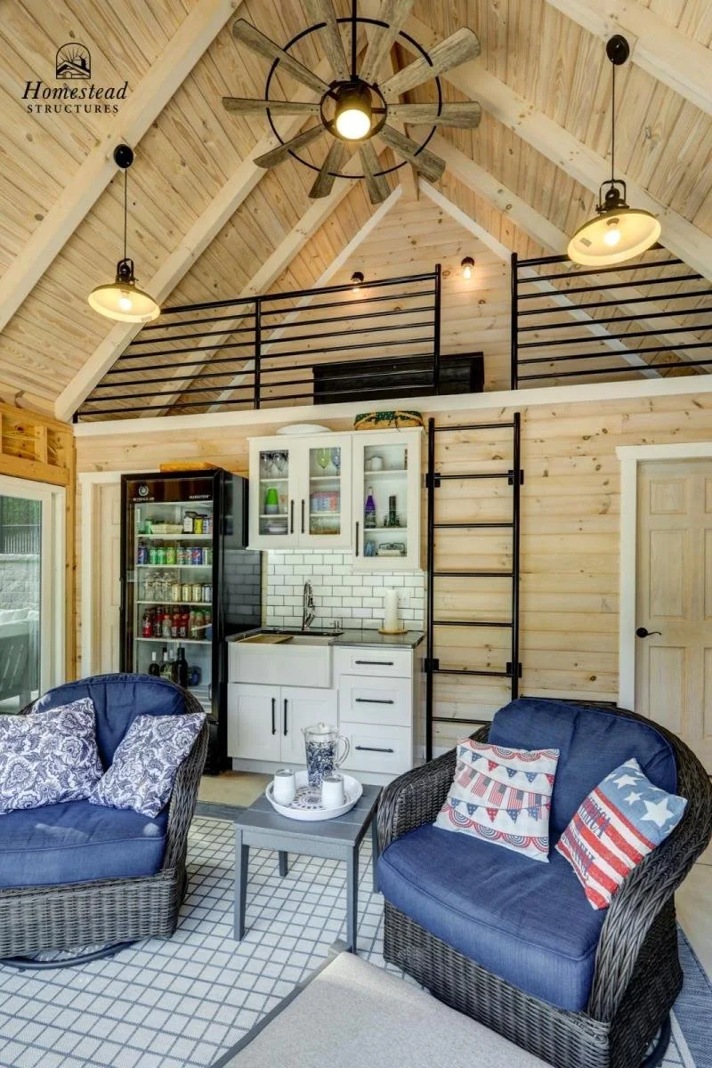 Interior of a rustic, wooden cabin living space with a loft area, ceiling fan, and hanging lights. There are two wicker chairs with patriotic-themed pillows, a small table with a pitcher and cups, a mini fridge, and a kitchenette with white cabinets 