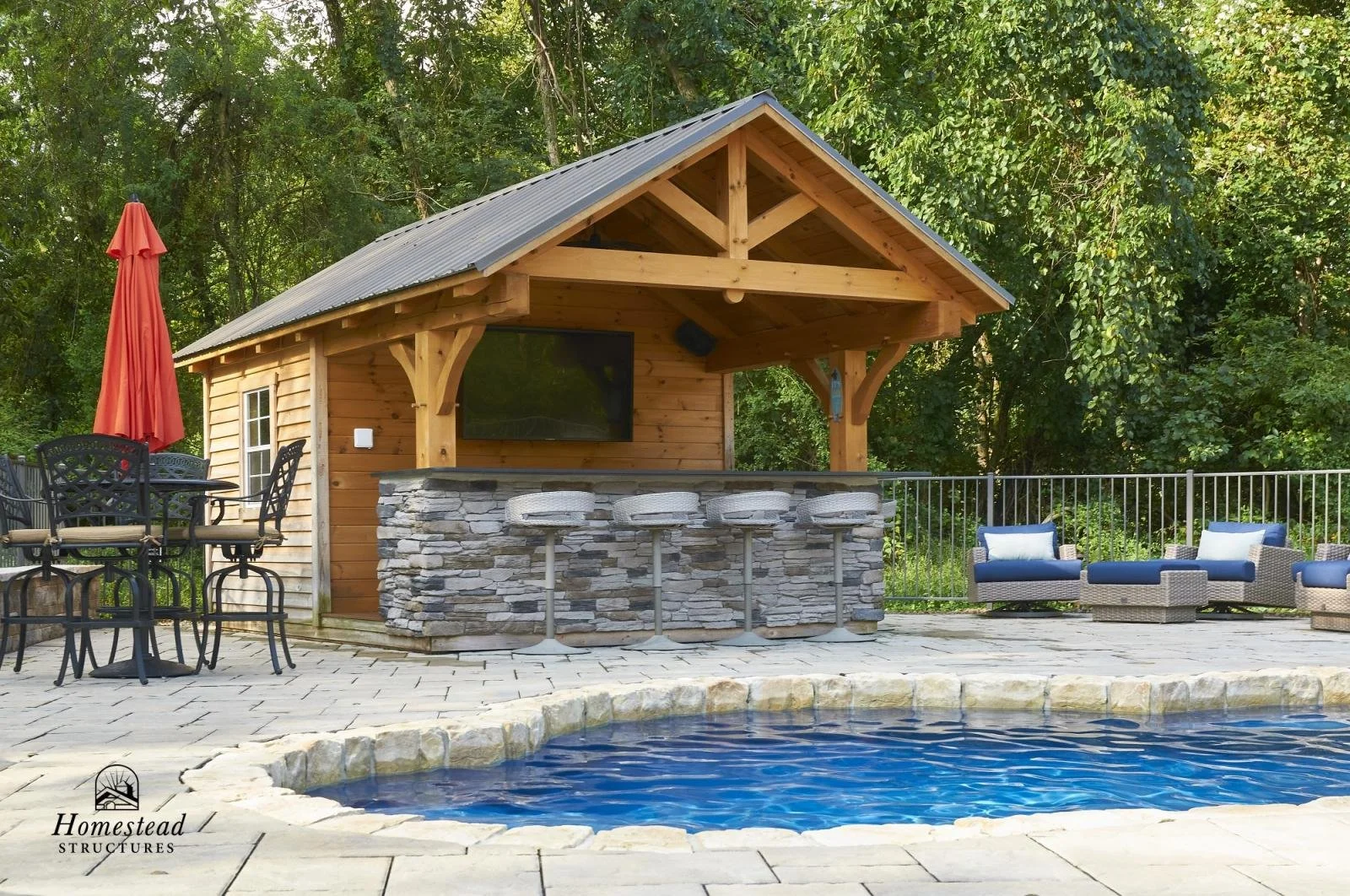 A backyard with a small wooden pool house, stone bar counter, bar stools, patio furniture, and a swimming pool, surrounded by trees.