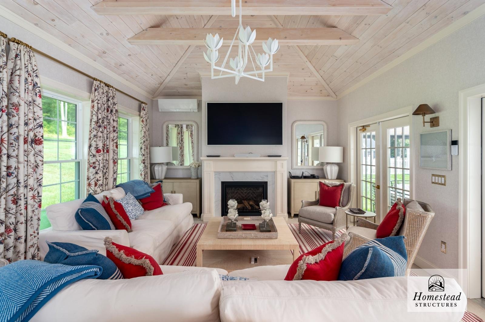 Living room with white sofas, armchairs, a stone fireplace, a large wall-mounted TV, windows with floral curtains, a wooden ceiling with rafters, and various lamps and decorative items.