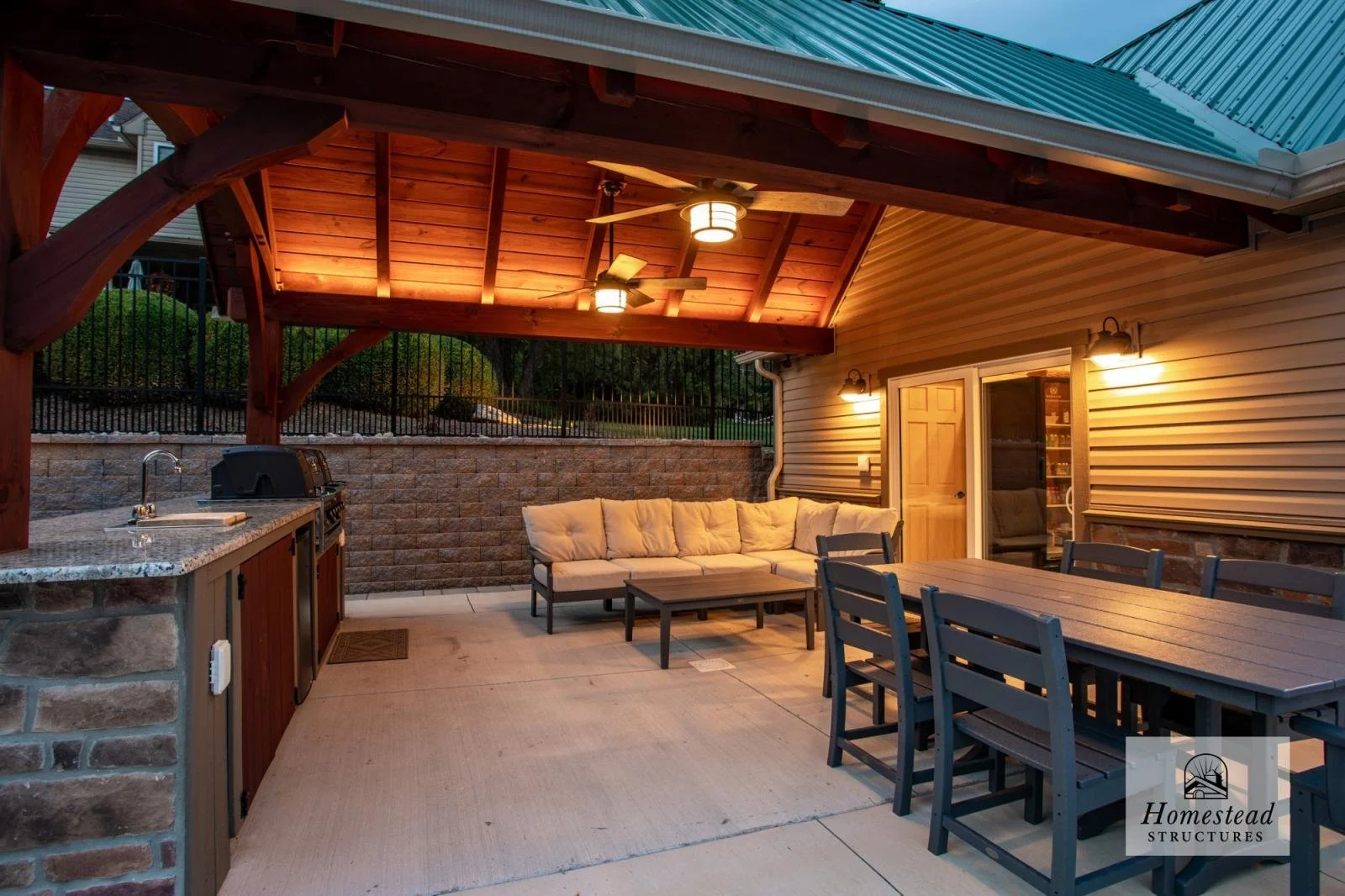 Covered outdoor patio with ceiling fans, lighting, a beige cushioned sofa, a wooden dining table with chairs, a small outdoor kitchen area with a sink and grill, and a sliding glass door leading into a house.