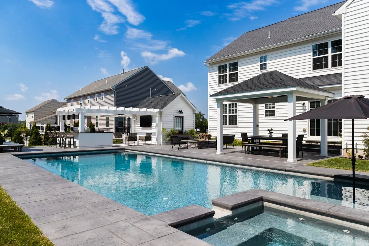 Residential backyard with a swimming pool, patio area with outdoor furniture, and a white house with a covered porch on a sunny day.