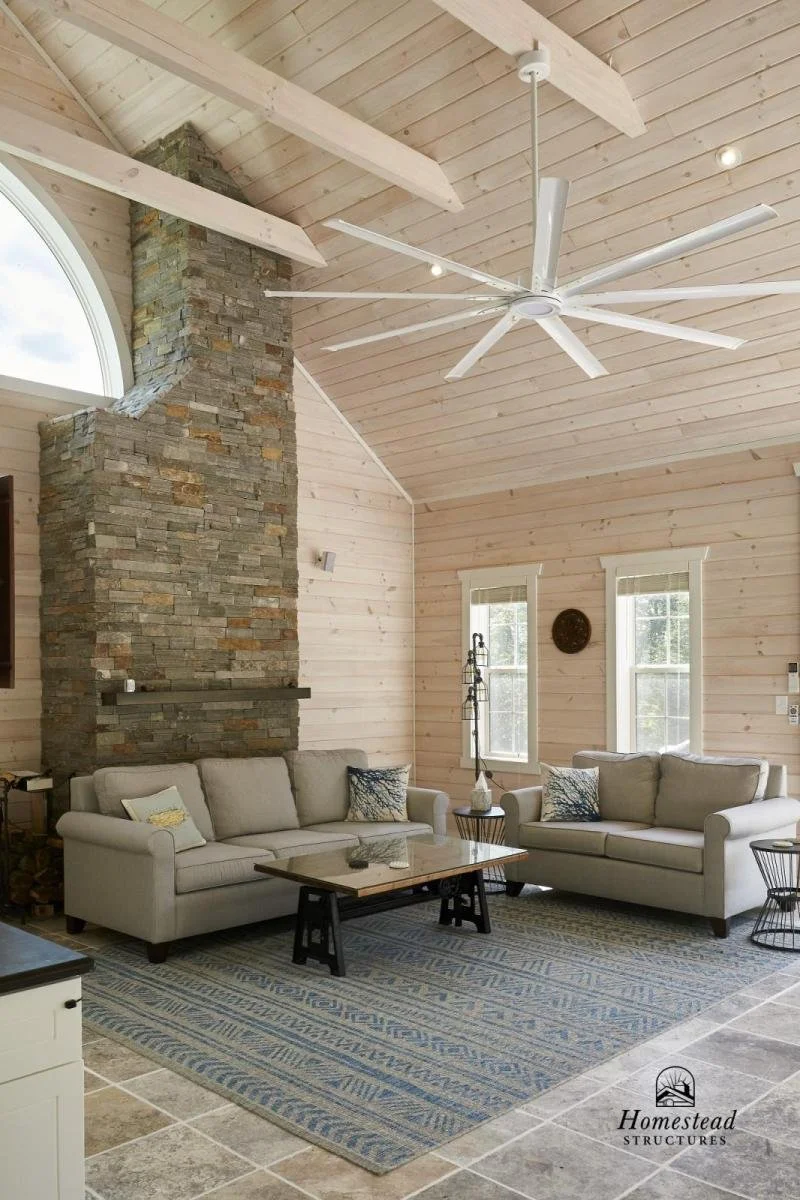 Living room with a high, wooden vaulted ceiling, large stone fireplace, light-colored sofas, patterned area rug, and two windows letting in natural light, with ceiling fan overhead.
