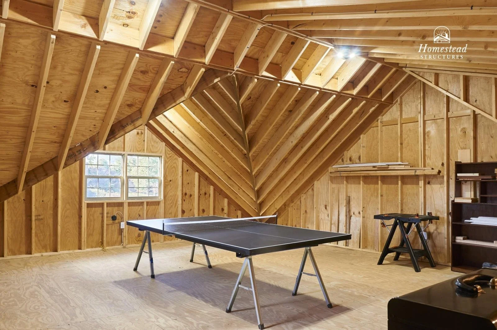 Unfinished wooden attic with a ping pong table, bookshelf, and a window, showing exposed wooden beams and walls.