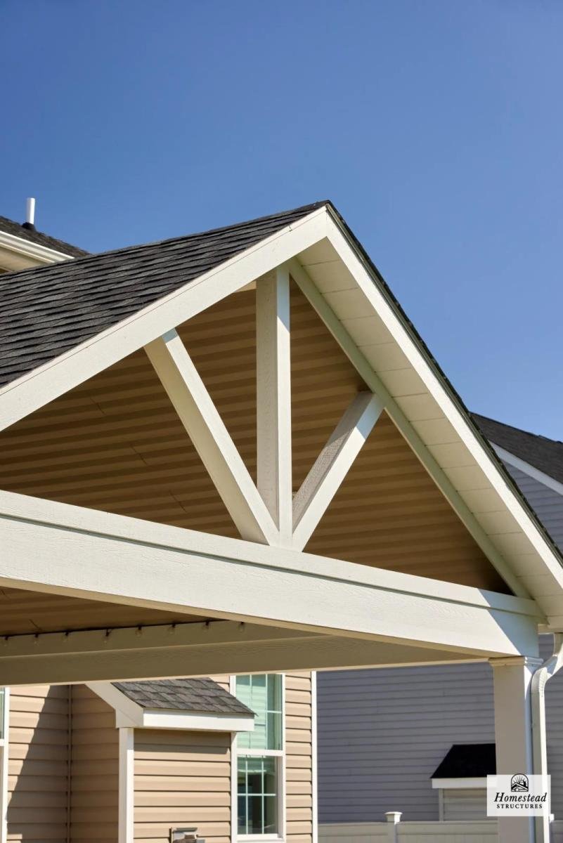 Close-up of a house's front gable with decorative exposed timber trusses painted white, beige siding, and a clear blue sky in the background.