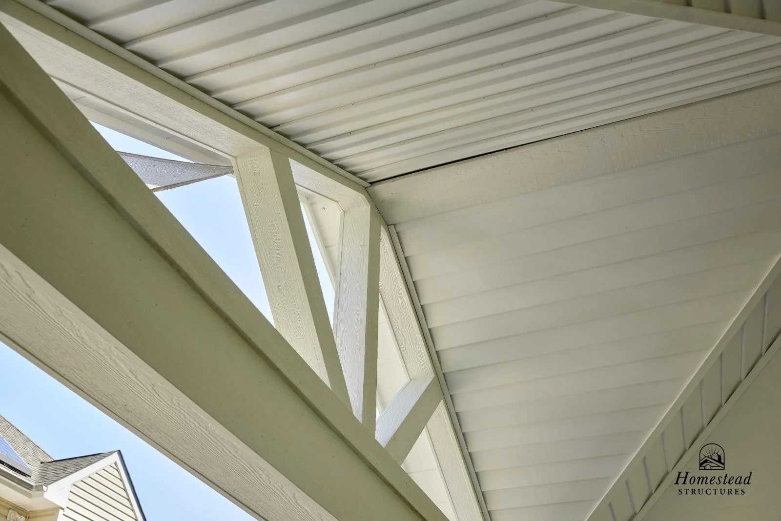 Close-up view of a house's porch ceiling and support beam with a partly open triangular attic vent, under a blue sky, with a small logo that reads 'Homestead Structures' in the bottom right corner.