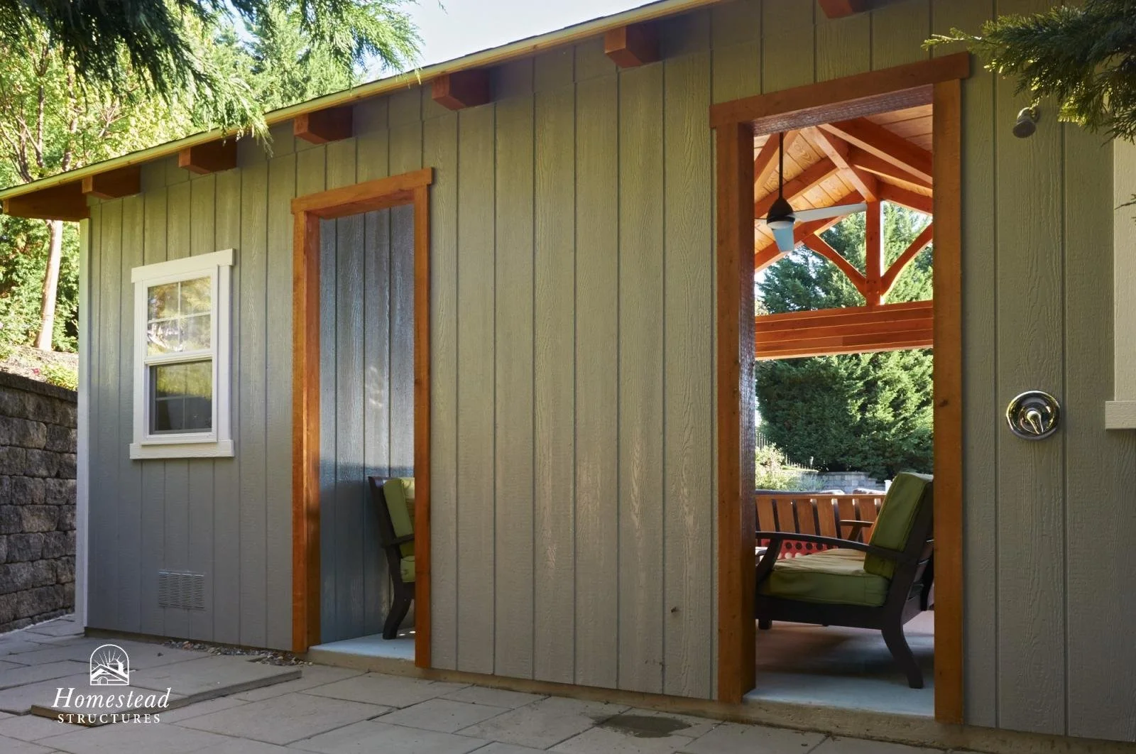 Exterior of a small, gray wooden shed with two doorways, one showing a covered porch with a ceiling fan and seating, and a window on the left. The shed has a stone path in front and trees in the background.