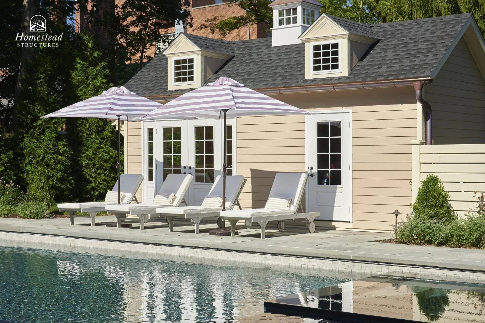 A small beige poolhouse with white trim, French doors, and dormer windows, situated next to a swimming pool with four white lounge chairs and two striped umbrellas on a concrete patio, surrounded by greenery.