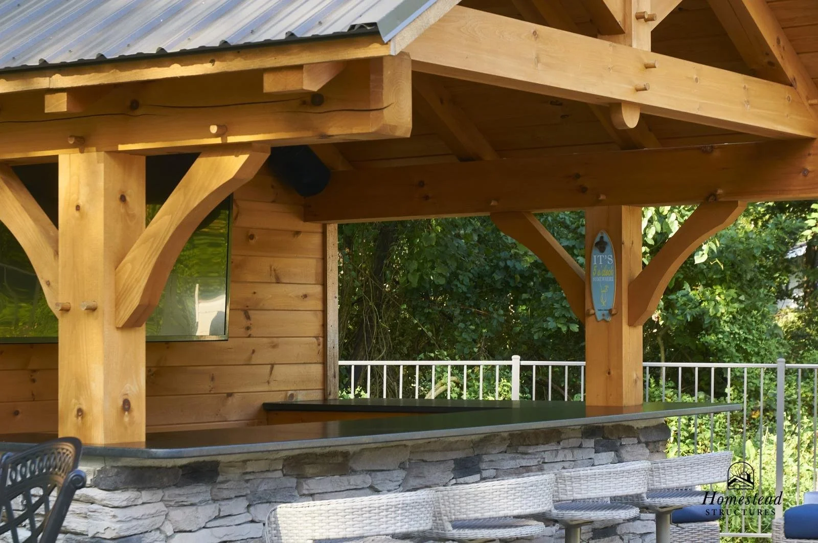 Outdoor bar area with wooden structure, stone countertop, and chairs, surrounded by trees and a white fence.