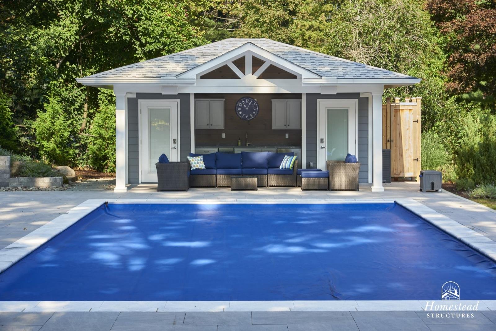 Backyard with a swimming pool covered by a blue safety cover, outdoor seating area with dark wicker sofa and chairs with blue cushions, small tables, and a small kitchen area in a pavilion, surrounded by trees.