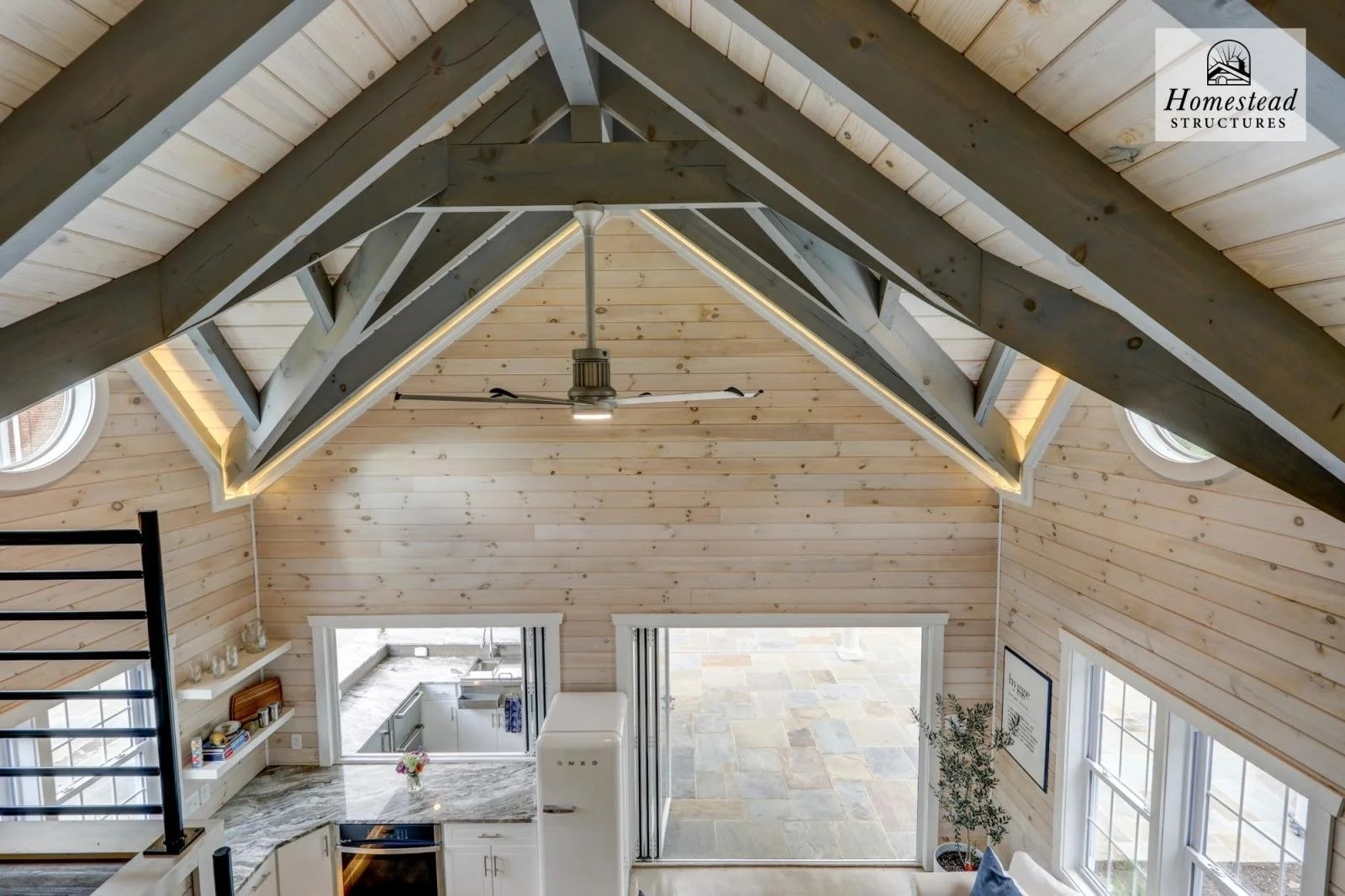 Interior view of a modern kitchen and living area with a vaulted ceiling, exposed beams, and large windows. The ceiling features wood paneling and a ceiling fan, with natural light flooding the space. The kitchen has white cabinetry and a marble coun