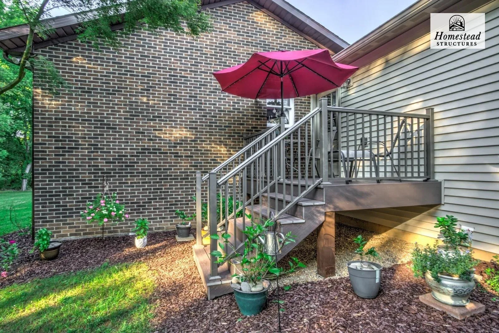 Backyard patio with wooden stairs, metal railing, potted plants, and a large pink patio umbrella next to a house with brick and vinyl siding.