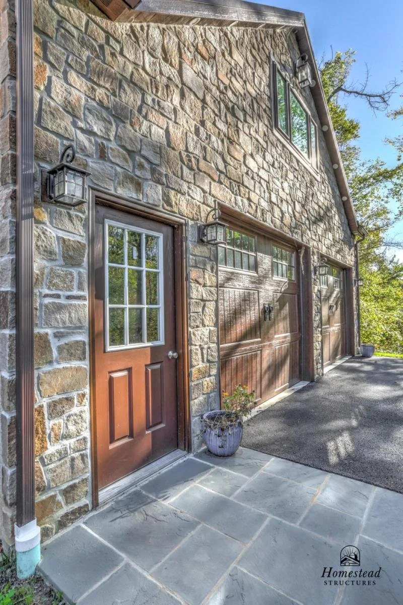 Exterior view of a stone house with two garage doors, a wooden front door, and outdoor wall lanterns, with a potted plant and a paved walkway.