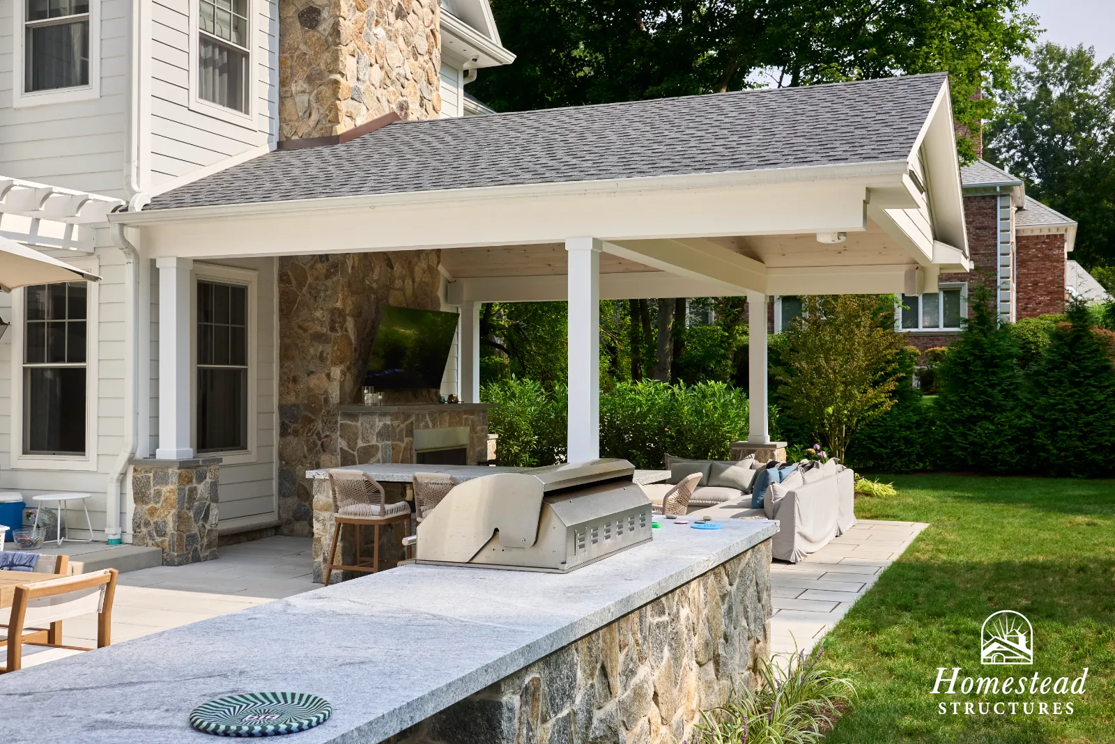 Backyard patio with outdoor seating area, stone fireplace, and a built-in grill under a roofed porch, surrounded by greenery.