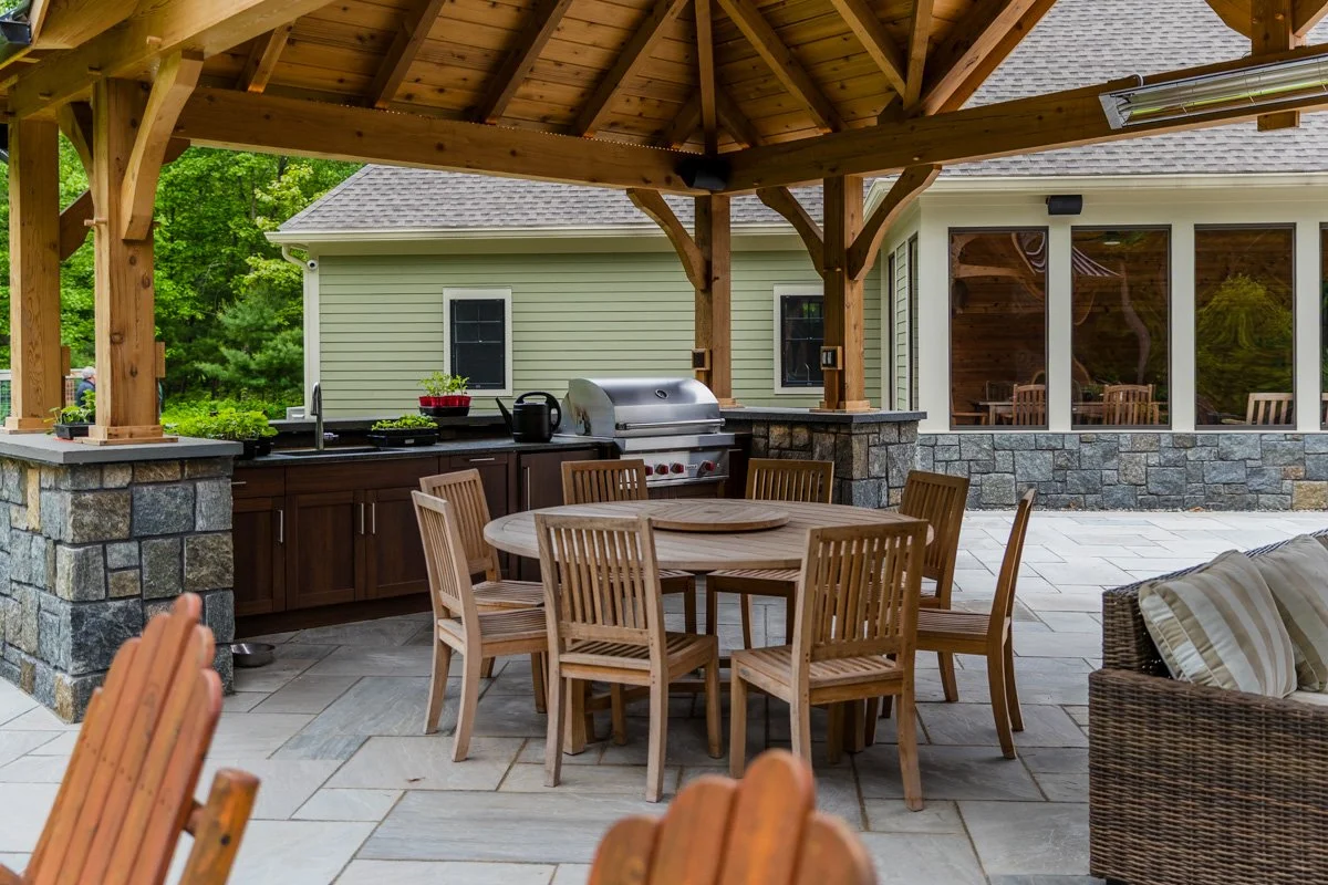 Outdoor patio with a round wooden table and six chairs, a built-in grill, and a view of a house with large glass windows, surrounded by greenery.