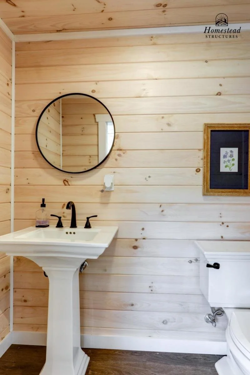 Bathroom with a pedestal sink, black faucet, round mirror, framed picture of purple flowers, white toilet, and wood-paneled walls.