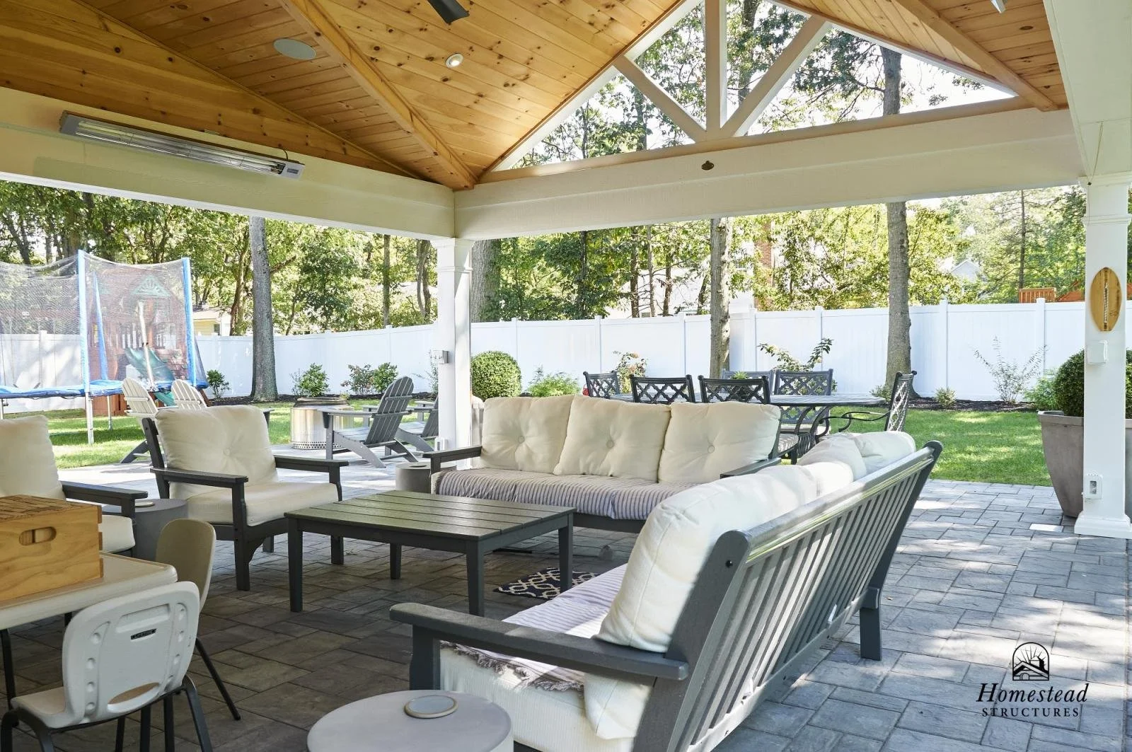 Covered outdoor patio with white cushioned seating, a wooden table, and surrounding greenery, including trees and a white fence.