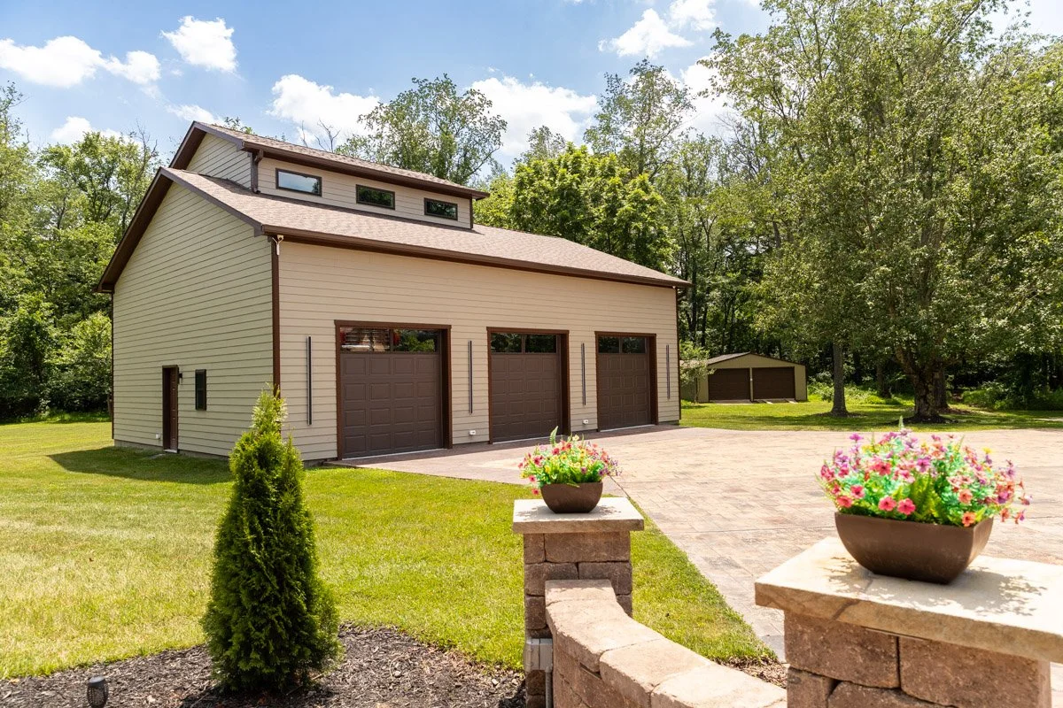 A large tan garage with three brown doors situated on a well-maintained lawn with a small shrub and flower pots, surrounded by trees under a partly cloudy sky.