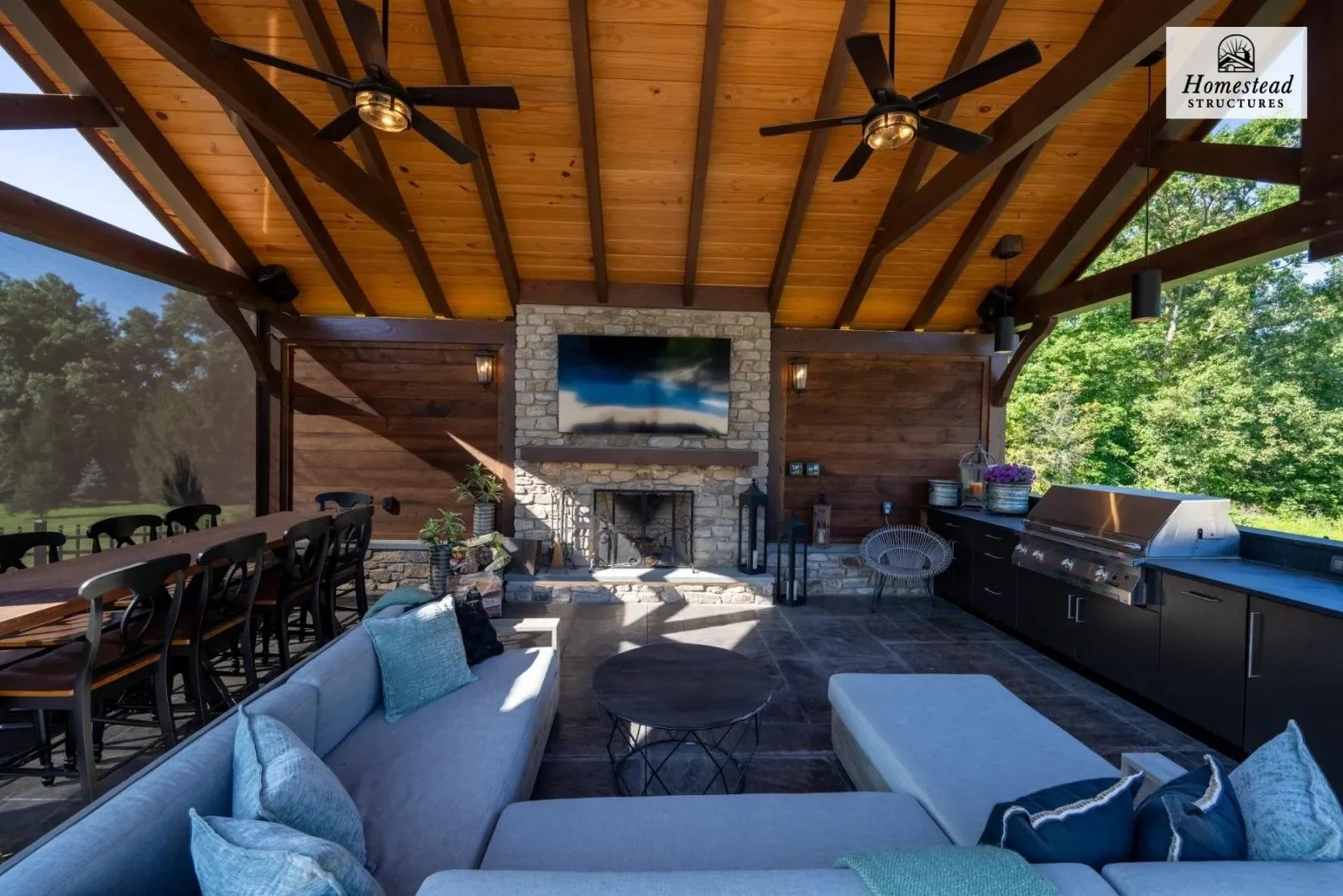 An outdoor living space with a covered wooden ceiling, ceiling fans, a stone fireplace with a TV mounted above, surrounded by stone and wood walls. There is a long dining table with chairs on the left, and a seating area with a sectional sofa, pillow
