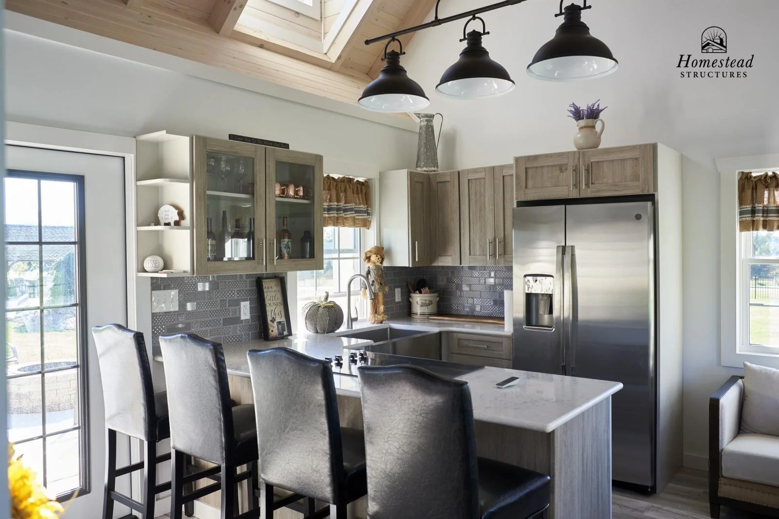 Modern kitchen with wooden cabinets, stainless steel refrigerator, island with black chairs, black pendant lights, and decorative items including a pumpkin and a pitcher with lavender.