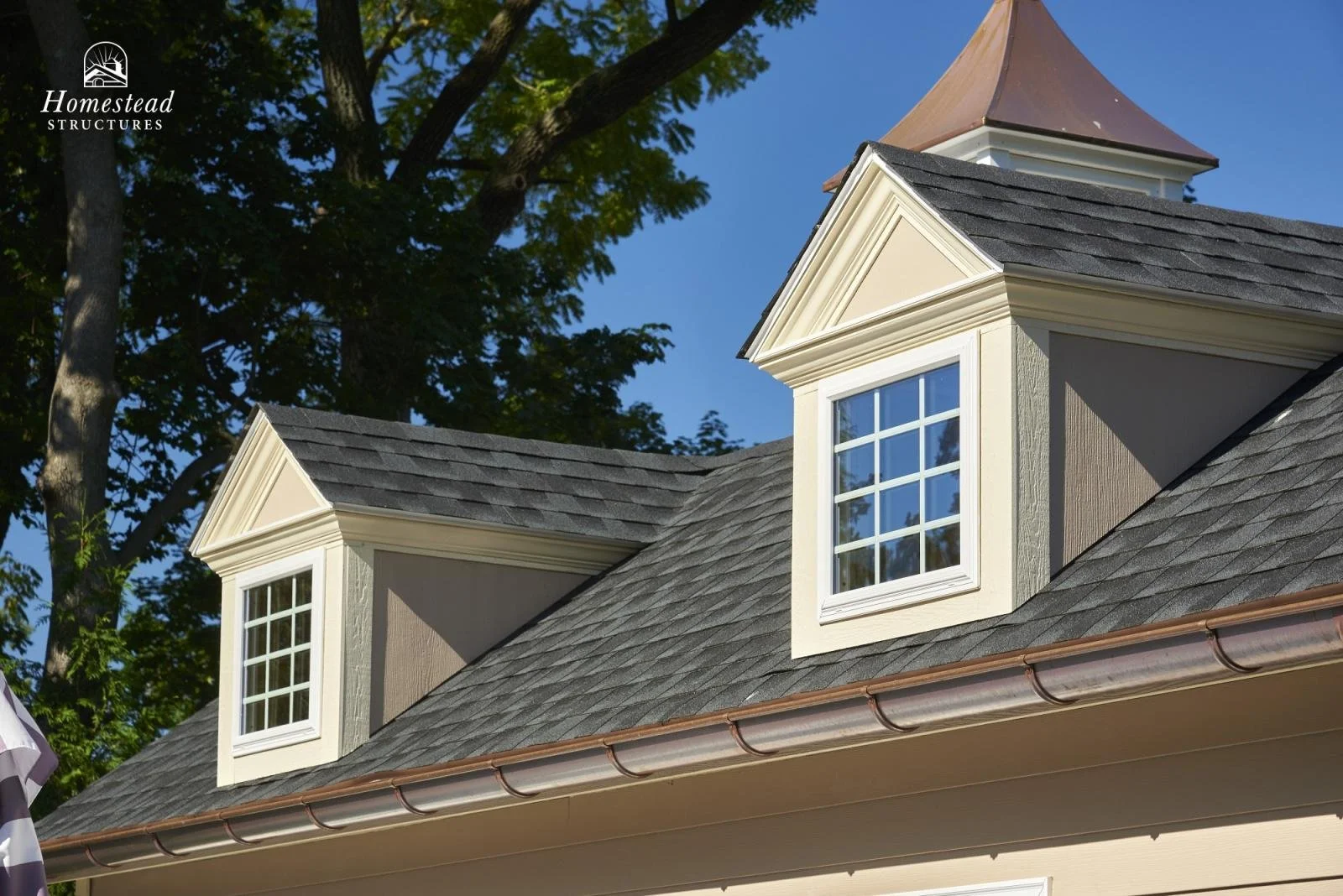 Close-up of a house's dormer windows with decorative molding on the roof, shingles, and a copper gutter, with trees and a blue sky in the background.