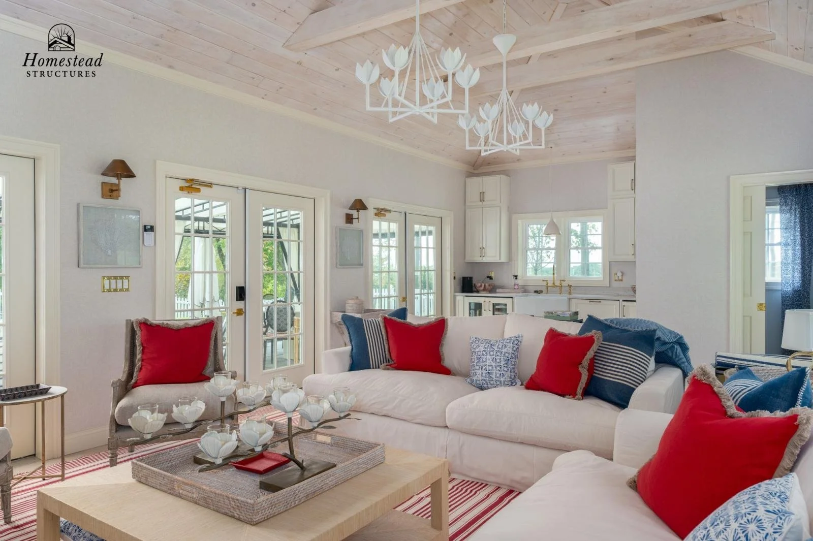 Living room with white sofas, red and blue pillows, a wooden coffee table with a decorative floral centerpiece, and glass doors leading to an outdoor area. The ceiling is wood-paneled with a white chandelier, and the kitchen with white cabinets is vi