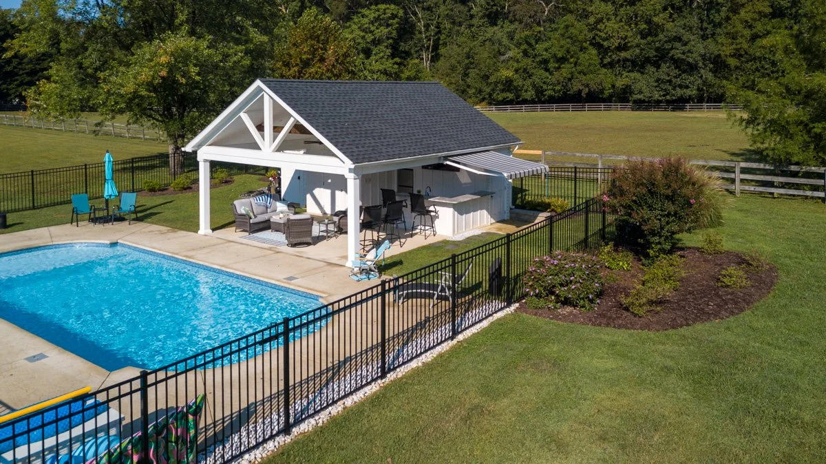 Backyard with swimming pool, poolside furniture, covered patio with seating and bar, surrounded by a fence, with trees and grass in the background.