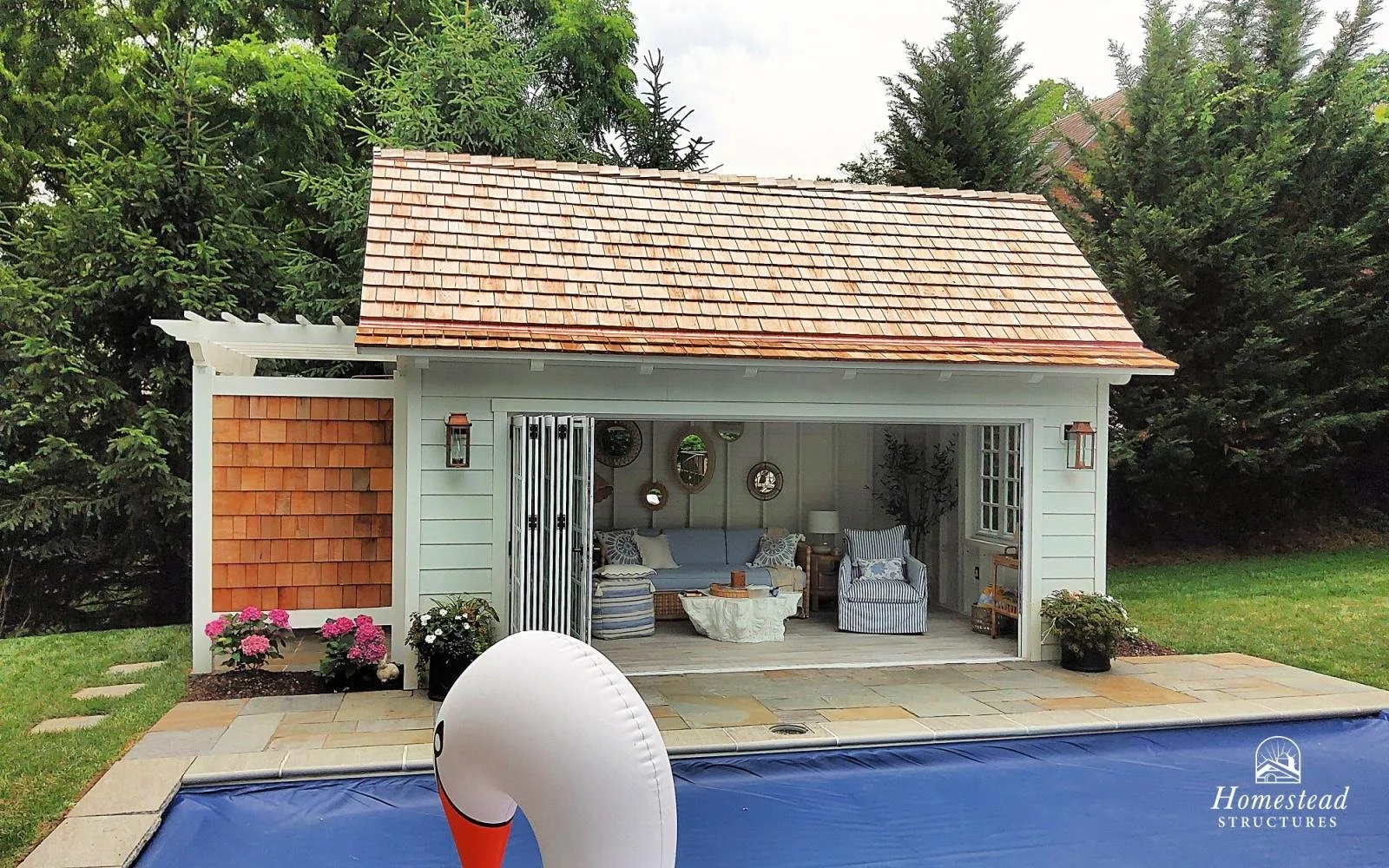 A small outdoor shed decorated as a cozy sitting area, situated next to a pool with a swan-shaped float, surrounded by green trees and plants, with homestead structures branding.