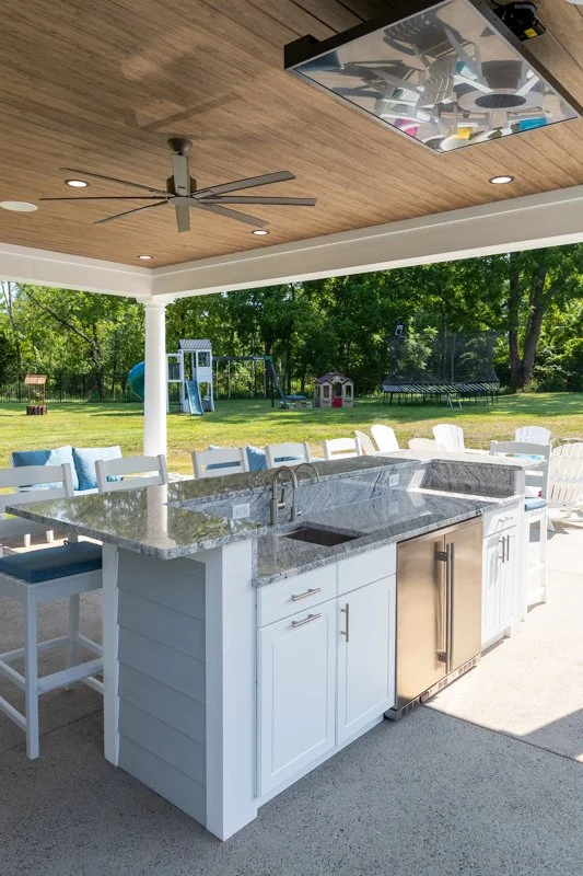 An outdoor kitchen with a gray granite countertop, white cabinets, and a stainless steel mini fridge. There are white chairs with blue cushions around the kitchen. In the background, there is a backyard with playground equipment and a trampoline, sur