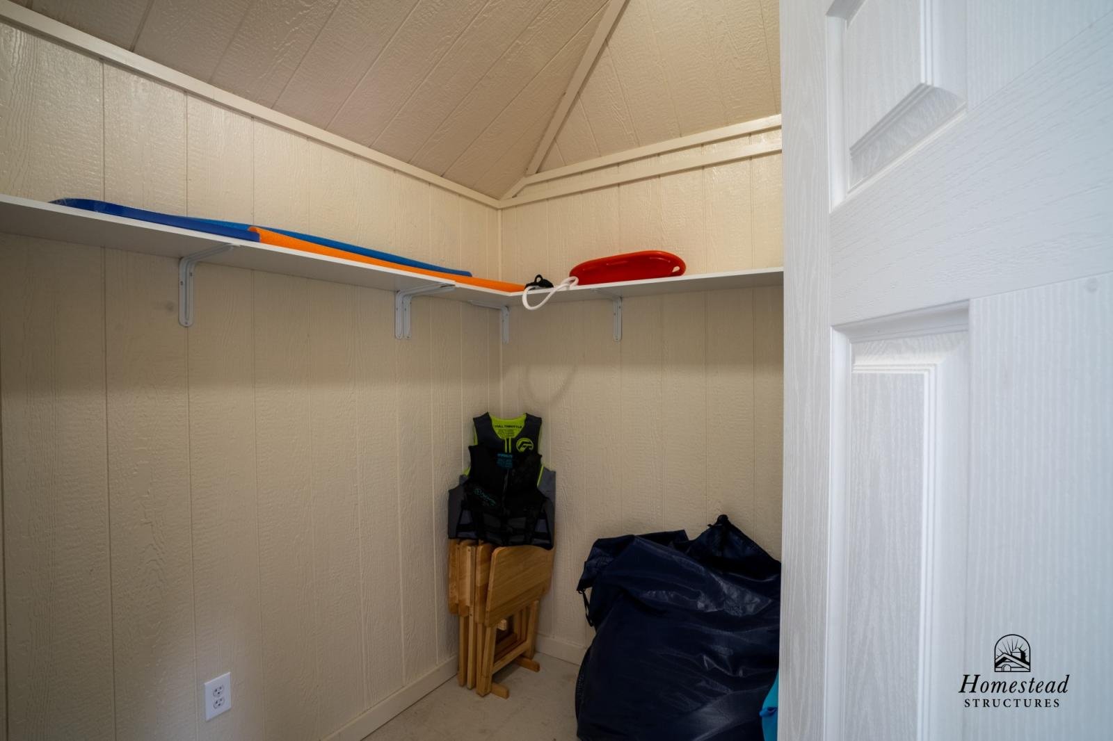 Small storage closet with beige paneled walls, white shelf holding bodyboards, red frisbee, and a cord. Folded wooden chairs and a black bag are on the floor.