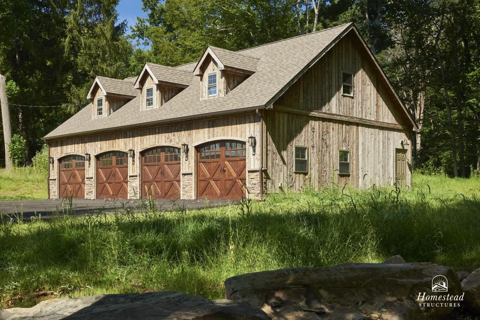A rustic barn with three garage doors, made of weathered wood, surrounded by green grass and trees.