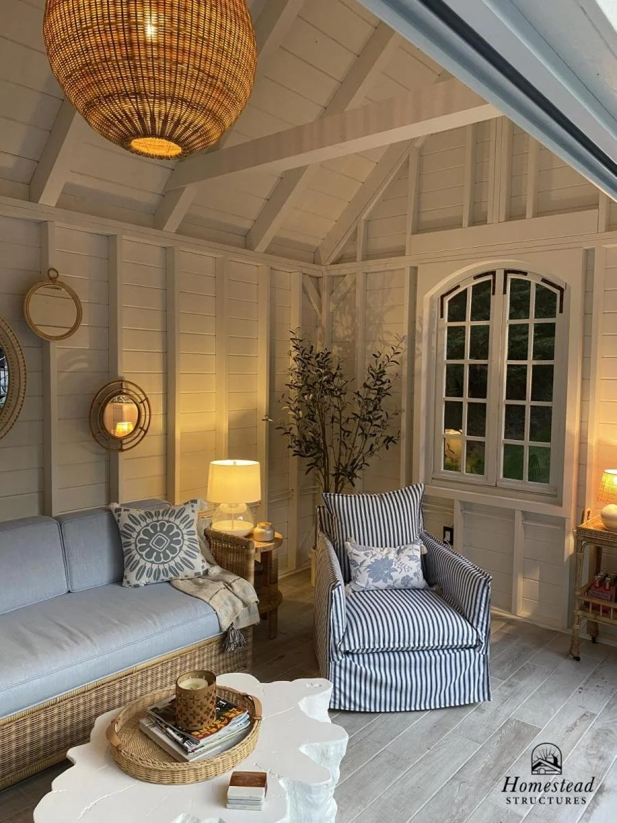 Cozy sunroom with white wooden walls and ceiling, wicker sofa with patterned cushion, striped armchair with floral cushion, side tables with lamps, a round white coffee table with candles and magazines, decorative mirrors on the wall, and a large arc