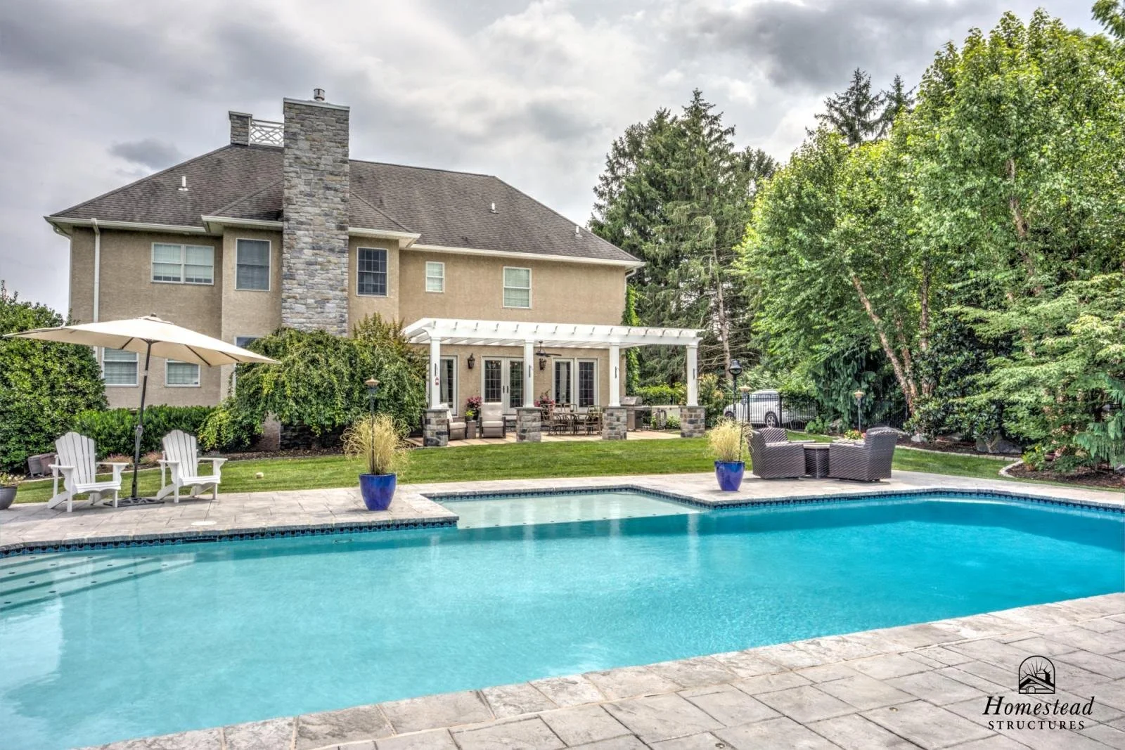 Backyard with a swimming pool, patio furniture, and a large house with a stone chimney, surrounded by trees and greenery.