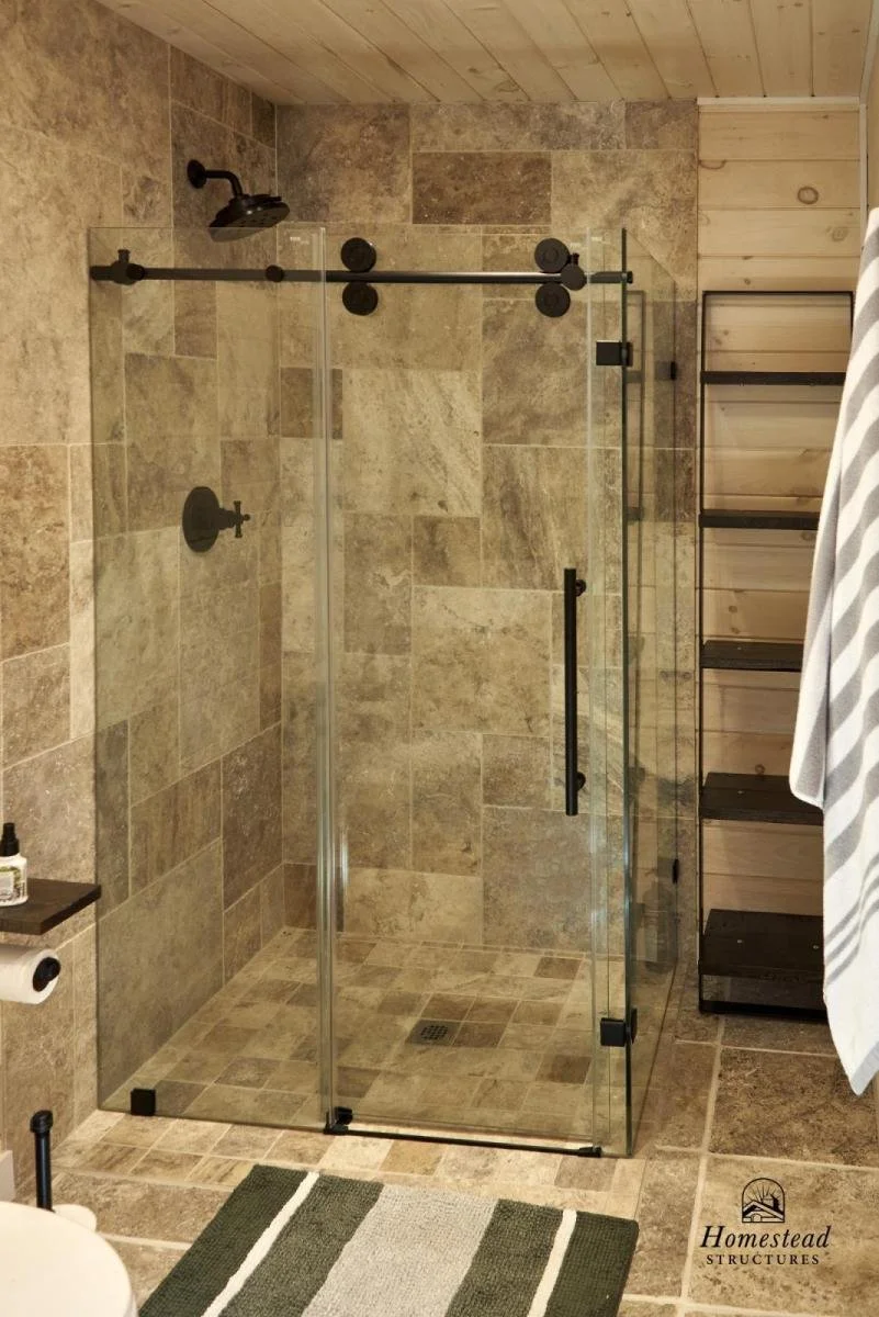 A walk-in shower with a black showerhead and handle, encased in glass with black hardware, in a rustic bathroom with tan stone tiles and a wooden wall on the right, alongside a black shelving unit and a white towel hanging.