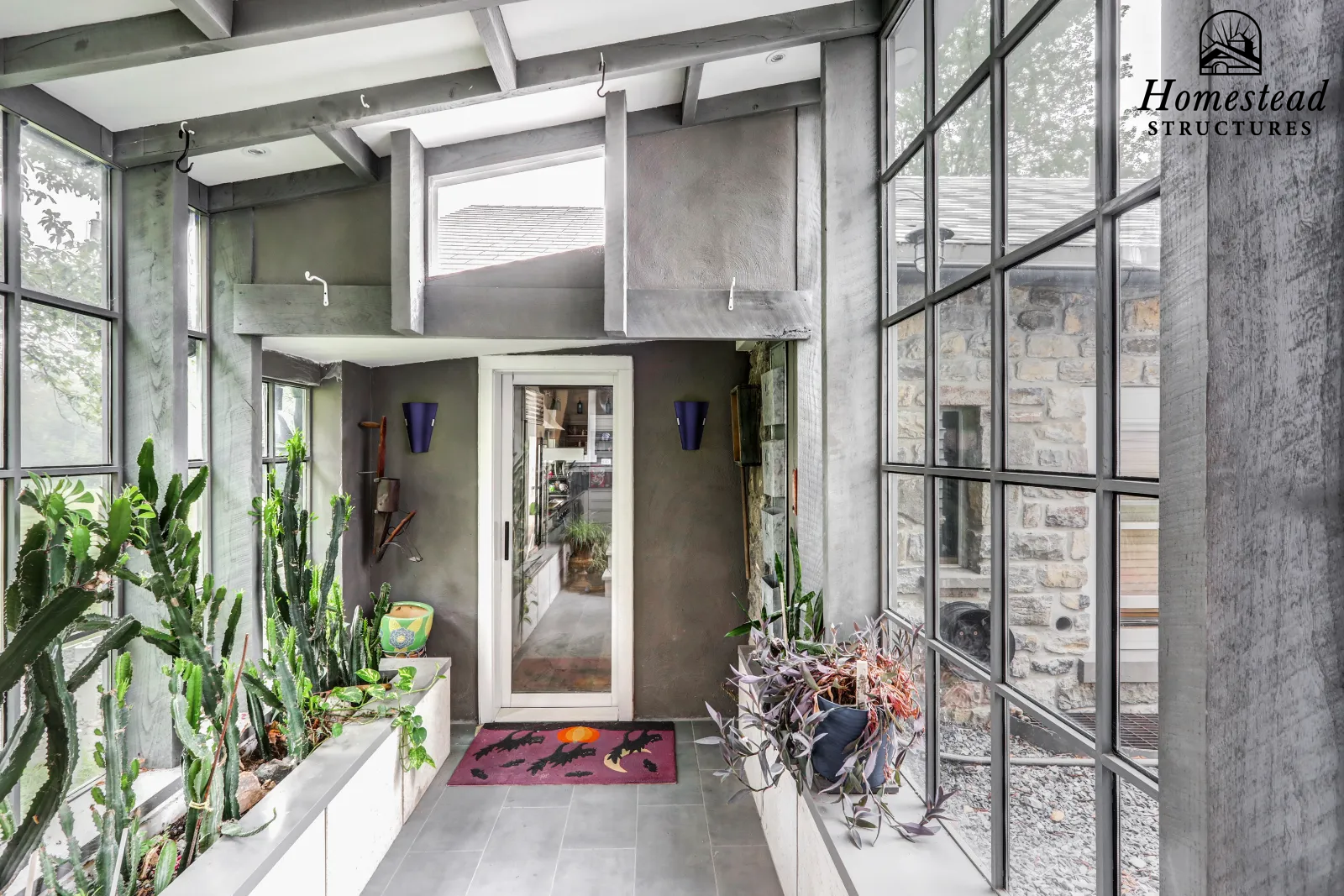 Sunroom with large windows, gray concrete walls, potted plants including cacti, a purple doormat with bats and a pumpkin, a glass door leading outside, and a view of neighboring houses with stone and wooden siding.