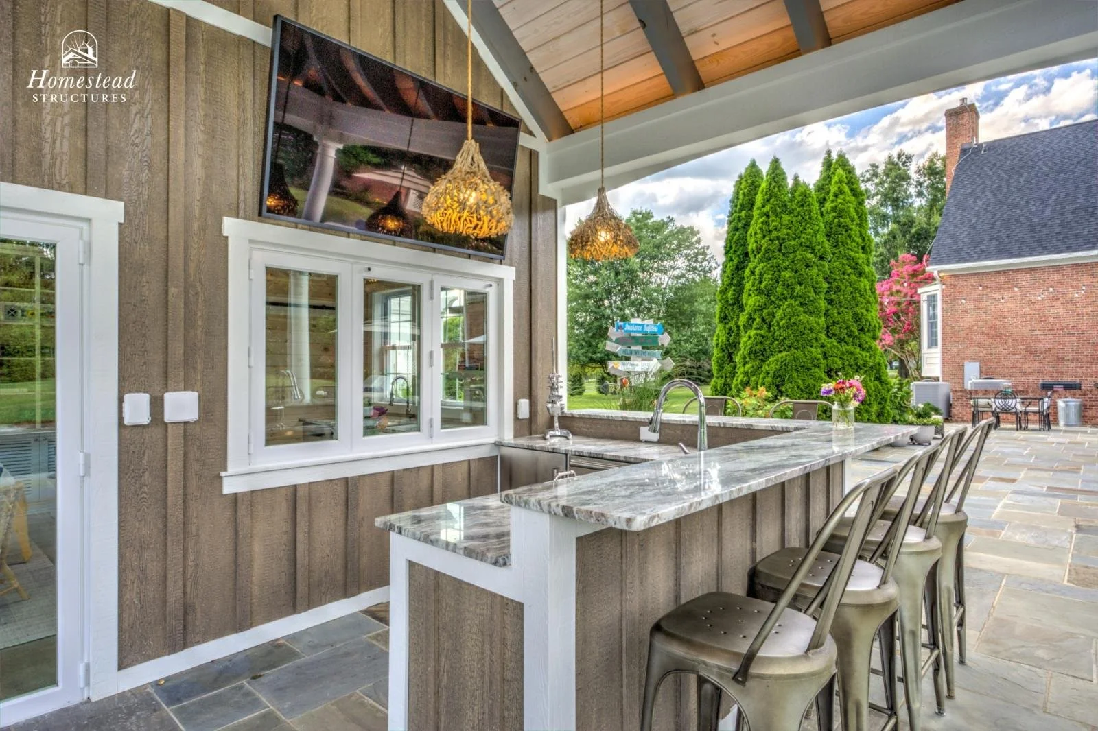 Outdoor kitchen with granite counter, three bar stools, and a view of tall evergreen trees and a brick house.