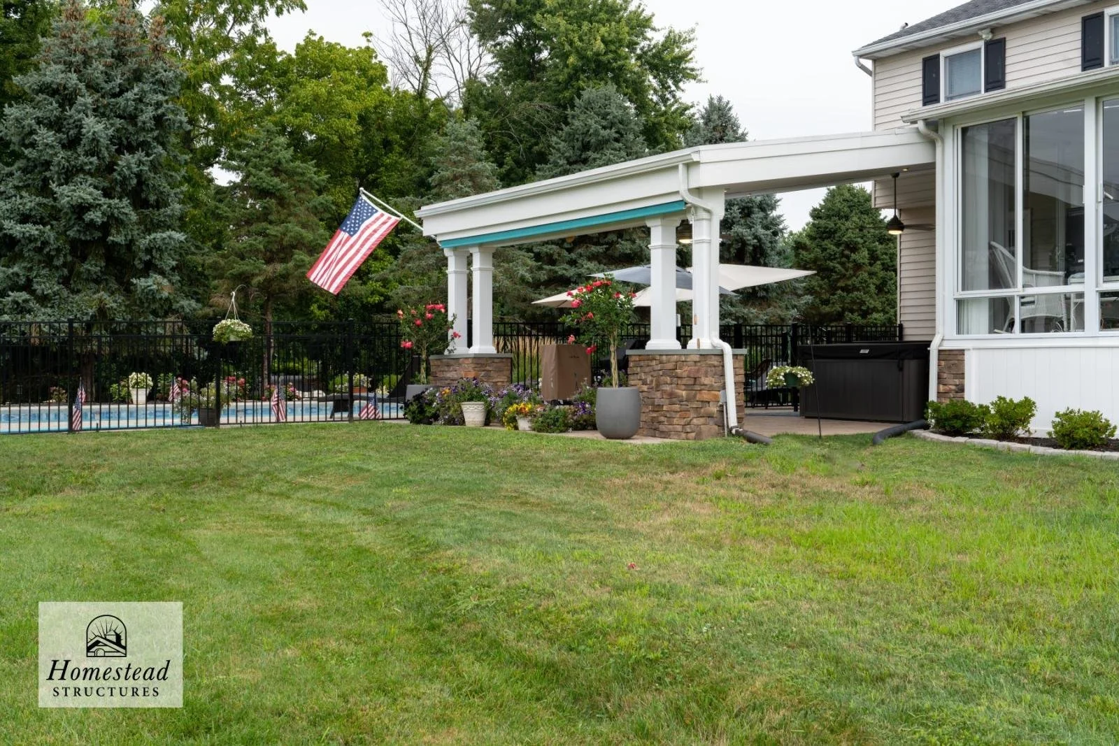 A backyard with a grassy lawn, a black metal fence, a swimming pool with an American flag, potted plants, flowers, and a covered patio with outdoor furniture, surrounded by trees.