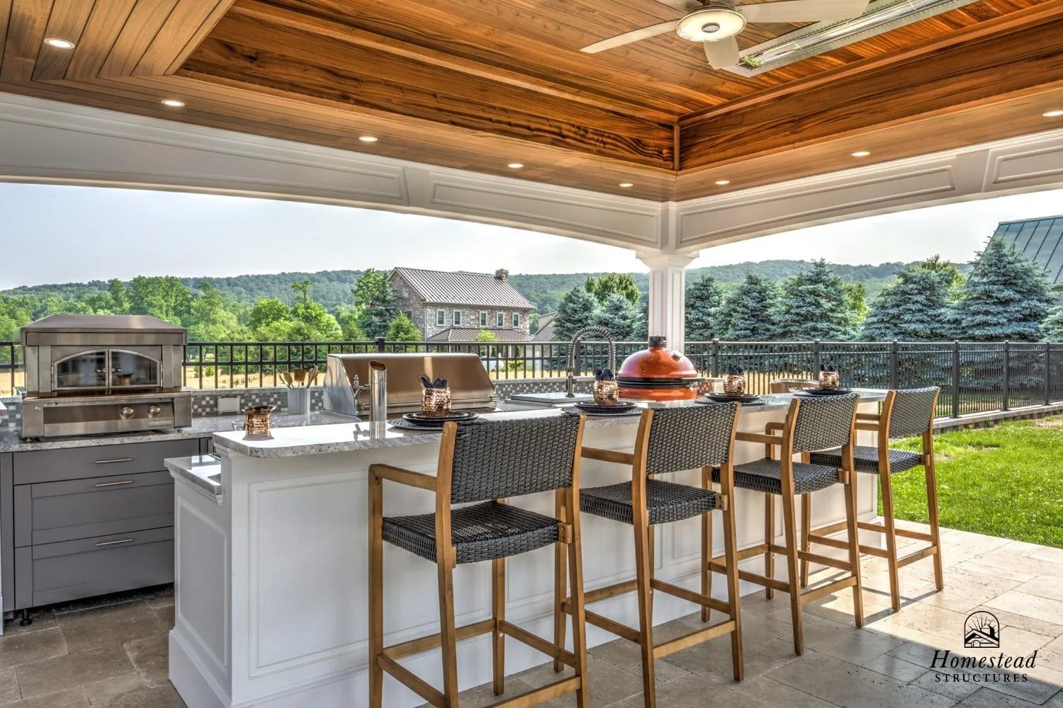 Outdoor kitchen with a countertop, grill, and bar seating, overlooking a yard with trees and neighborhood houses.