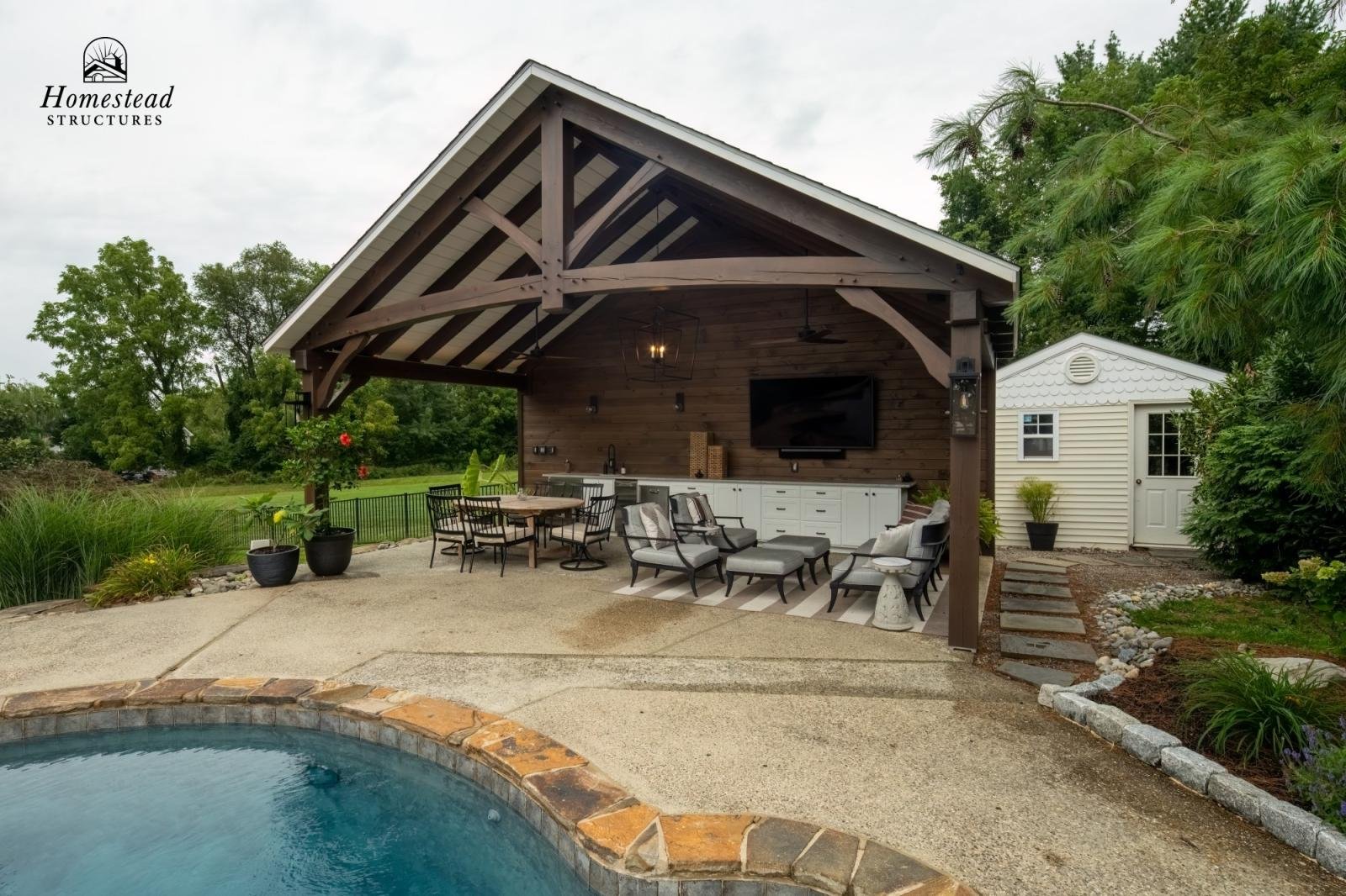Outdoor patio area featuring a covered pavilion with seating, a mountain-style shed, a swimming pool in the foreground, and lush green trees in the background.