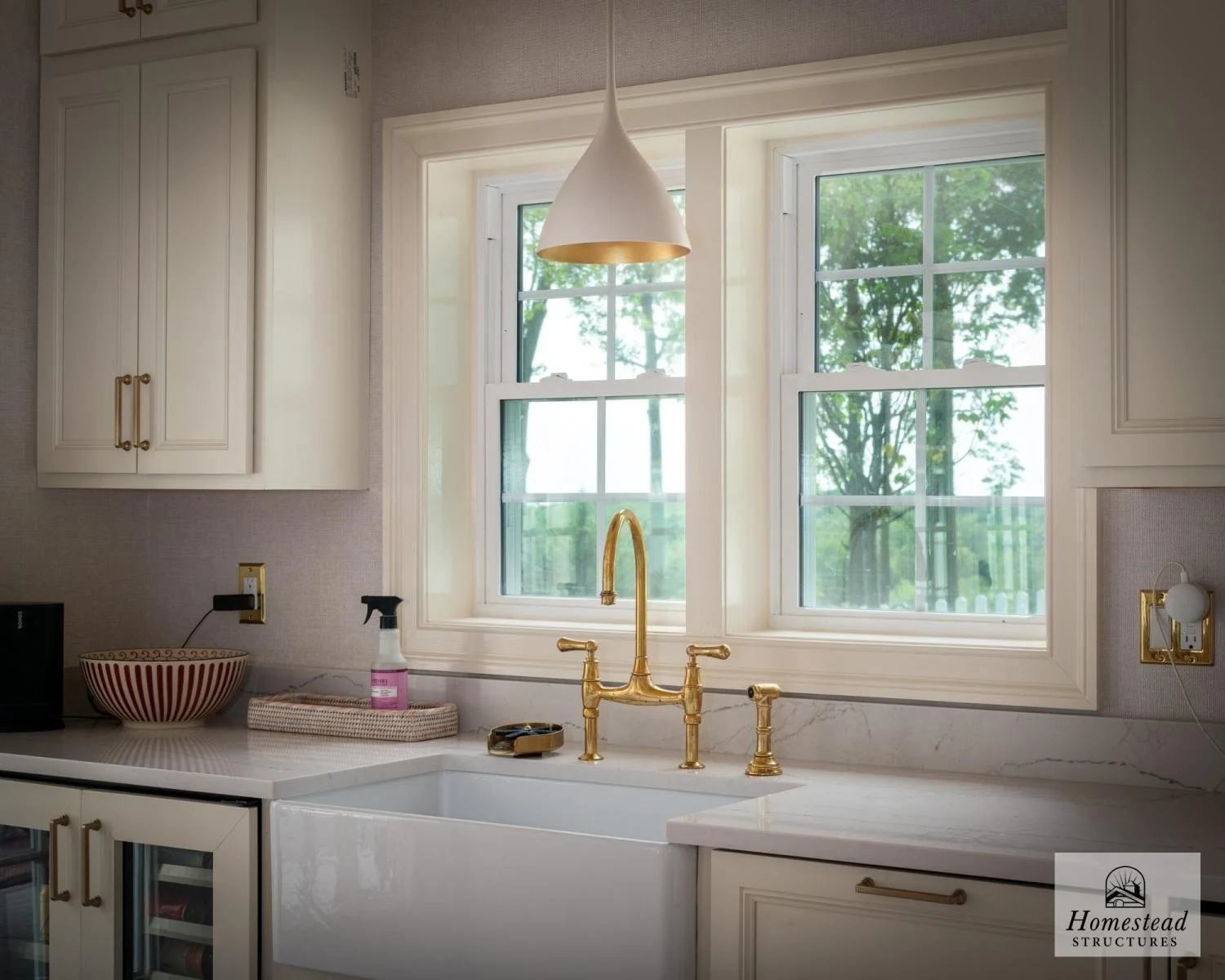 A kitchen with white cabinetry, a white farmhouse sink, and brass fixtures under a large window with a view of green trees outside. There is a pendant light hanging above the sink and a bowl on the counter.