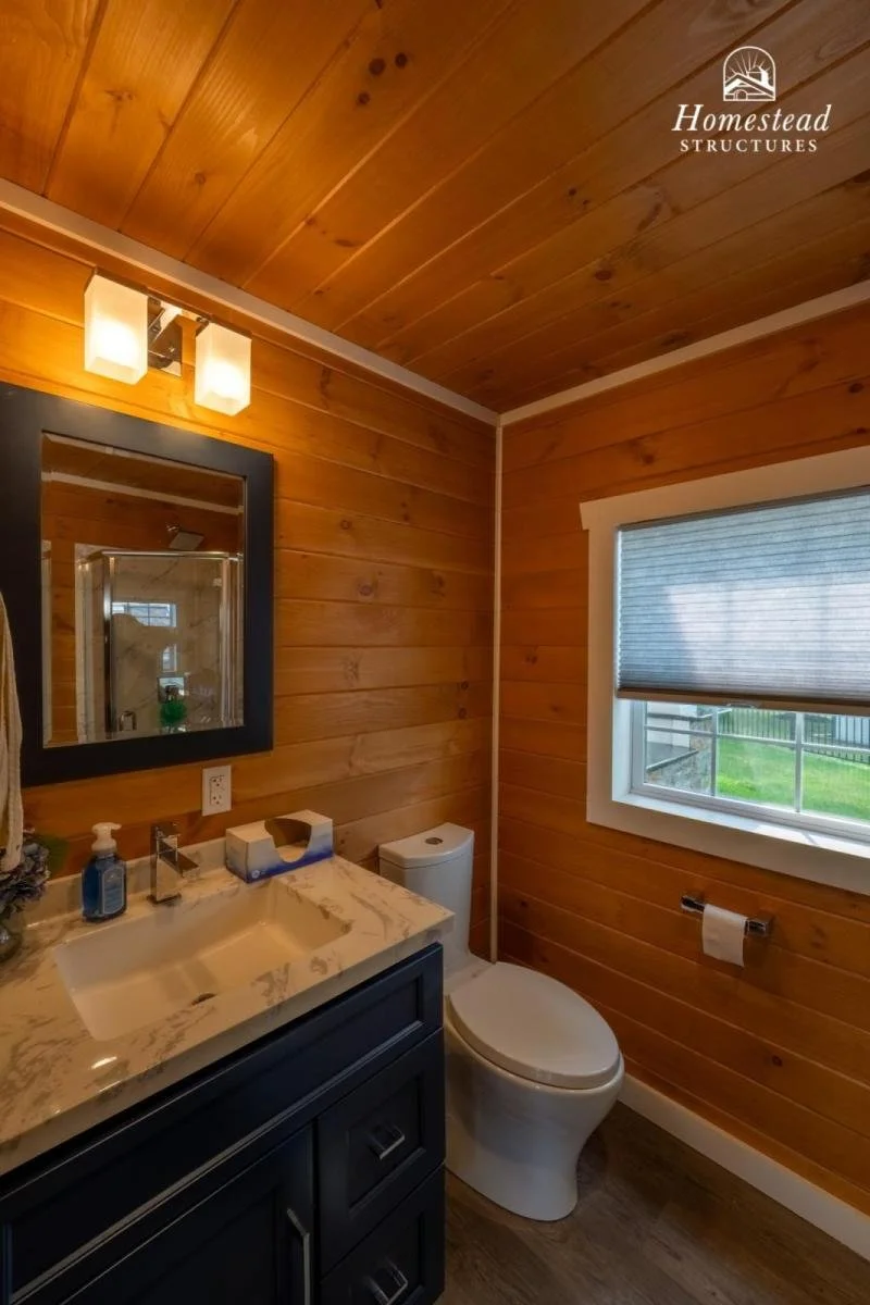 A small bathroom with wooden panel walls and ceiling. It has a white sink with a black cabinet, a mirror, a window with a shade, a toilet, and a bathroom light fixture. The logo of Homestead Structures is in the top right corner.