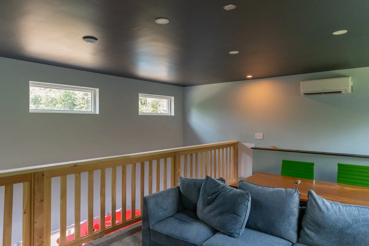A living room with a grey couch, green chairs, a wooden table, and a wooden railing. There are three small horizontal windows and an air conditioning unit on the wall.