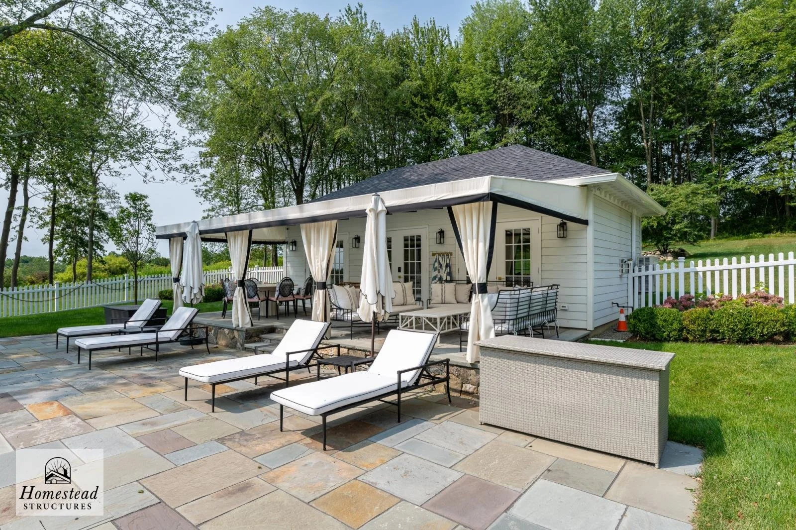 Backyard patio with lounge chairs, an outdoor seating area with curtains, and a small house surrounded by a white picket fence and trees.