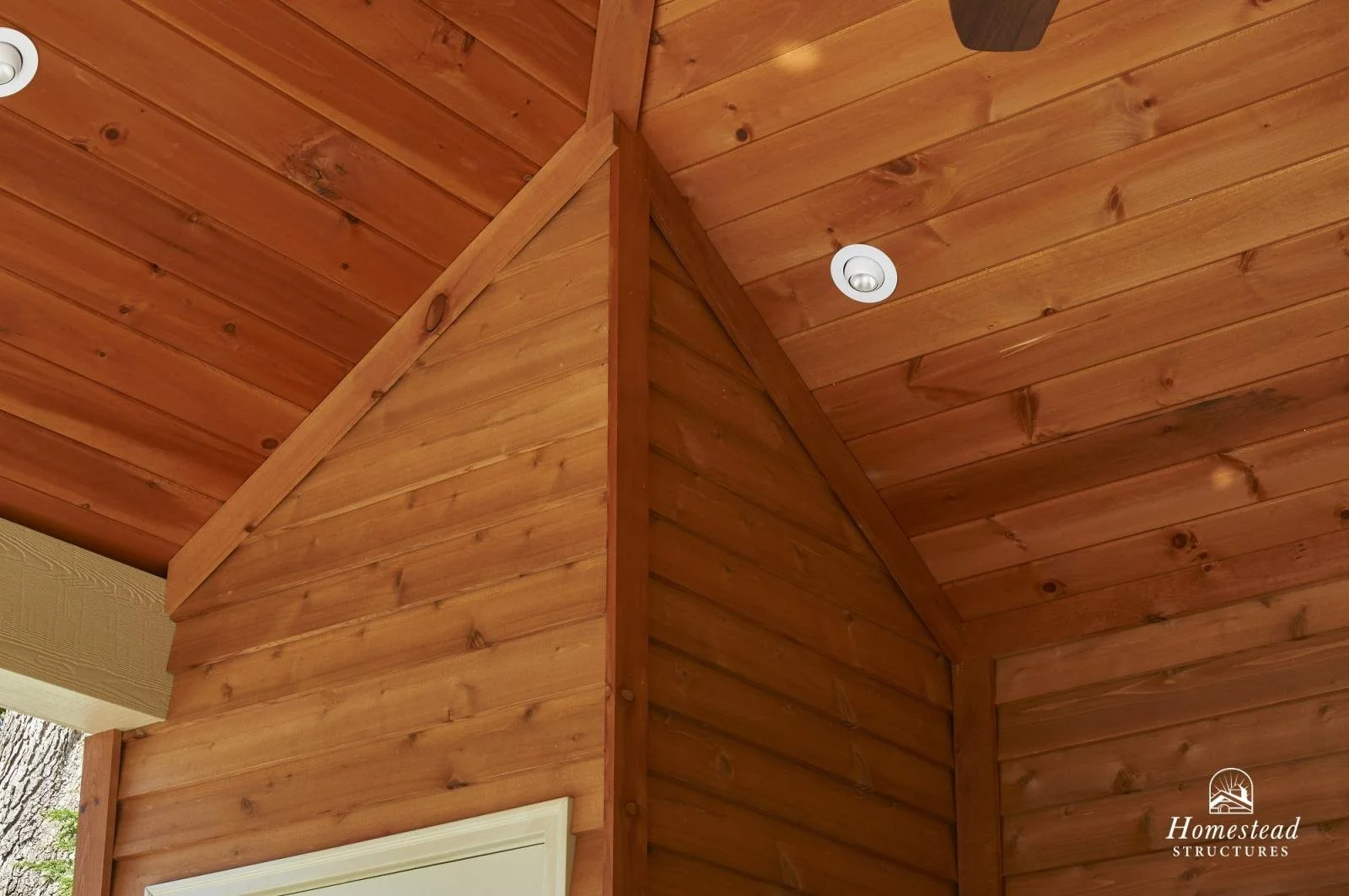 Wooden interior corner with cedar-paneled walls and ceiling, a small white circular ceiling light, and a white-framed window at the lower left corner.