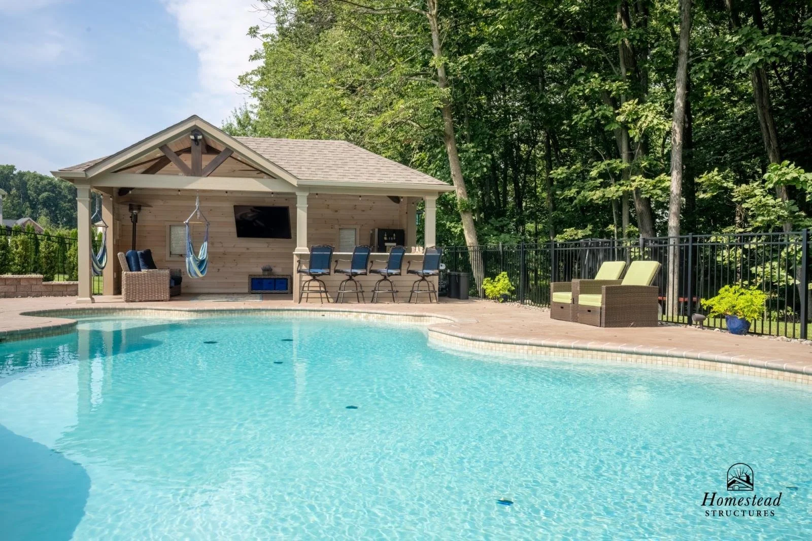 A backyard swimming pool with a poolside cabana and seating area, surrounded by lush green trees and a black metal fence.