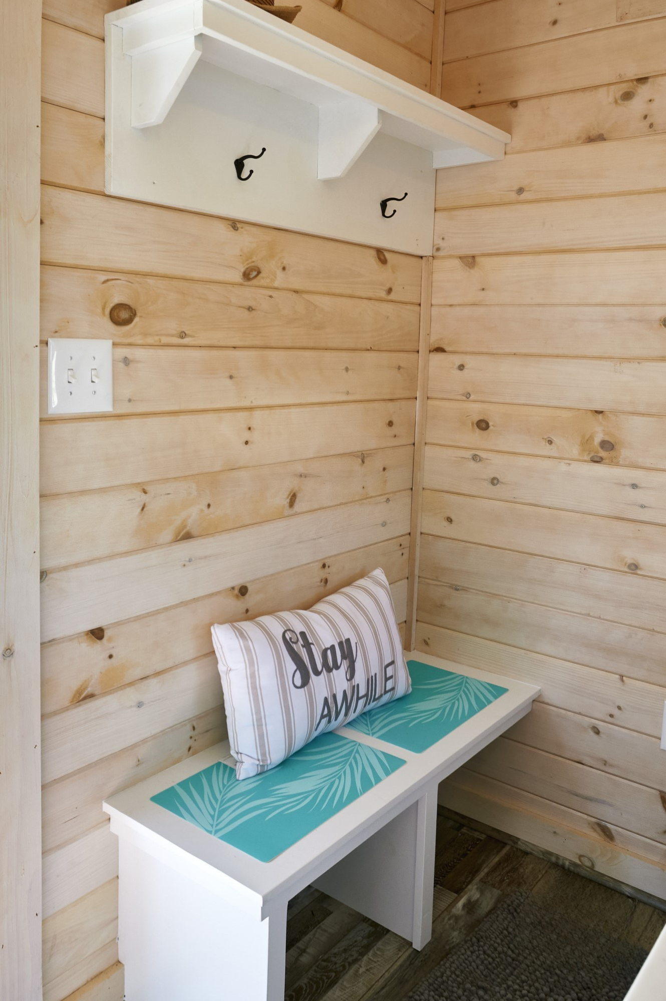 A small wooden entryway with a white bench, a striped pillow reading 'Stay Awhile,' and teal mats with palm leaf designs, against wood-paneled walls.