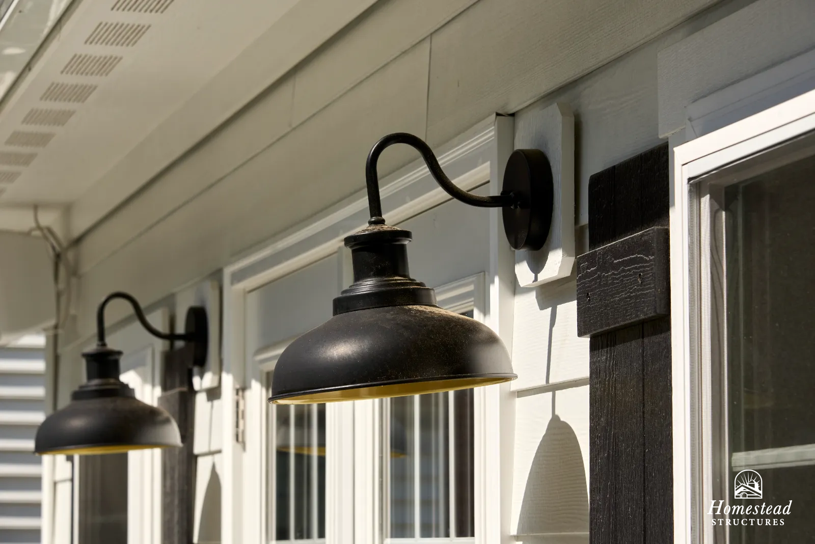 Two black outdoor wall sconces with yellow interior lining mounted on a light-colored house exterior with white-framed windows and black decorative accents.