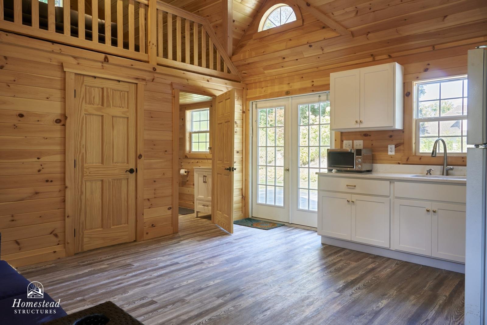 Interior of a wooden cabin kitchen with white cabinets, a microwave, a sink, and large windows and glass doors letting in natural light.