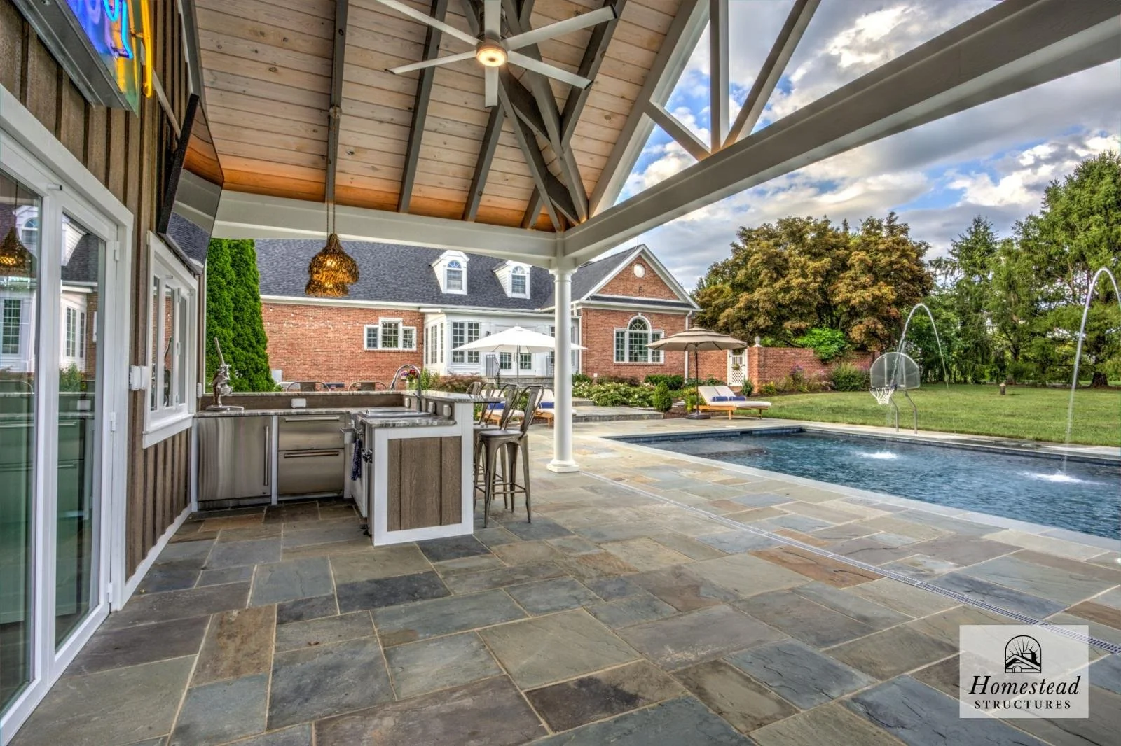 Outdoor covered patio area with a built-in kitchenette, bar stools, and a view of a swimming pool with fountains, lounge chairs, umbrellas, and a backyard with trees and grass.