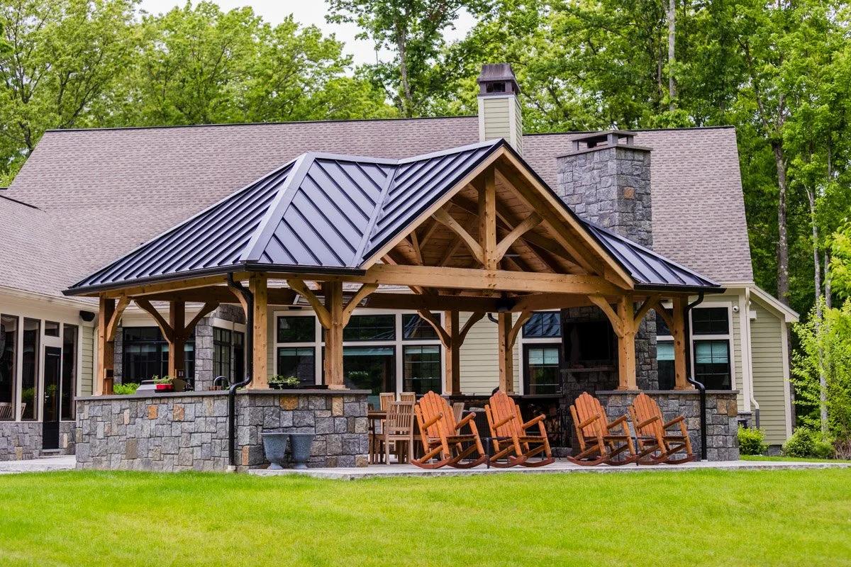 Backyard patio area with a covered wooden pergola, stone walls, outdoor furniture, and a barbecue grill, surrounded by green grass and trees.
